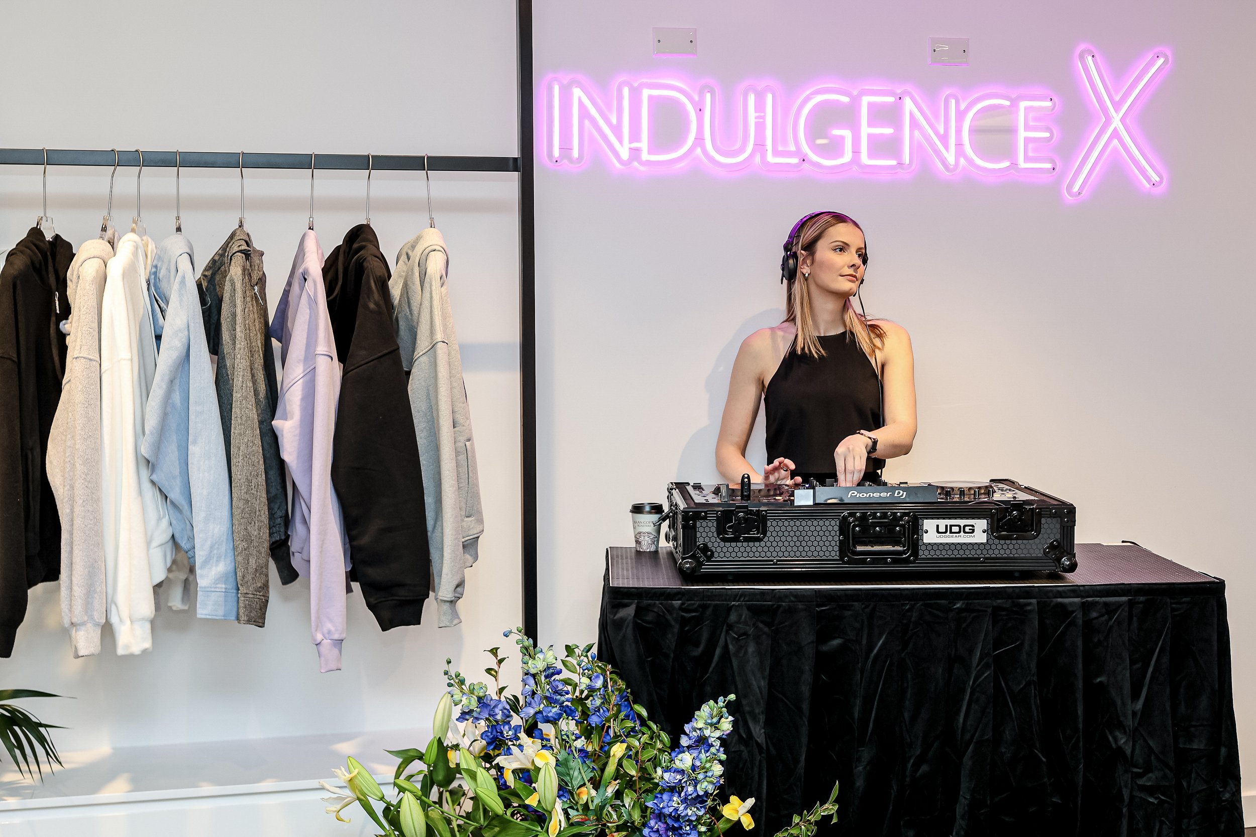 A woman DJ wearing headphones stands behind DJ equipment with a neon sign that reads 'Indulgence' and an 'X' on the wall. There are clothing racks with hoodies hanging on the left, and a floral arrangement in the foreground.