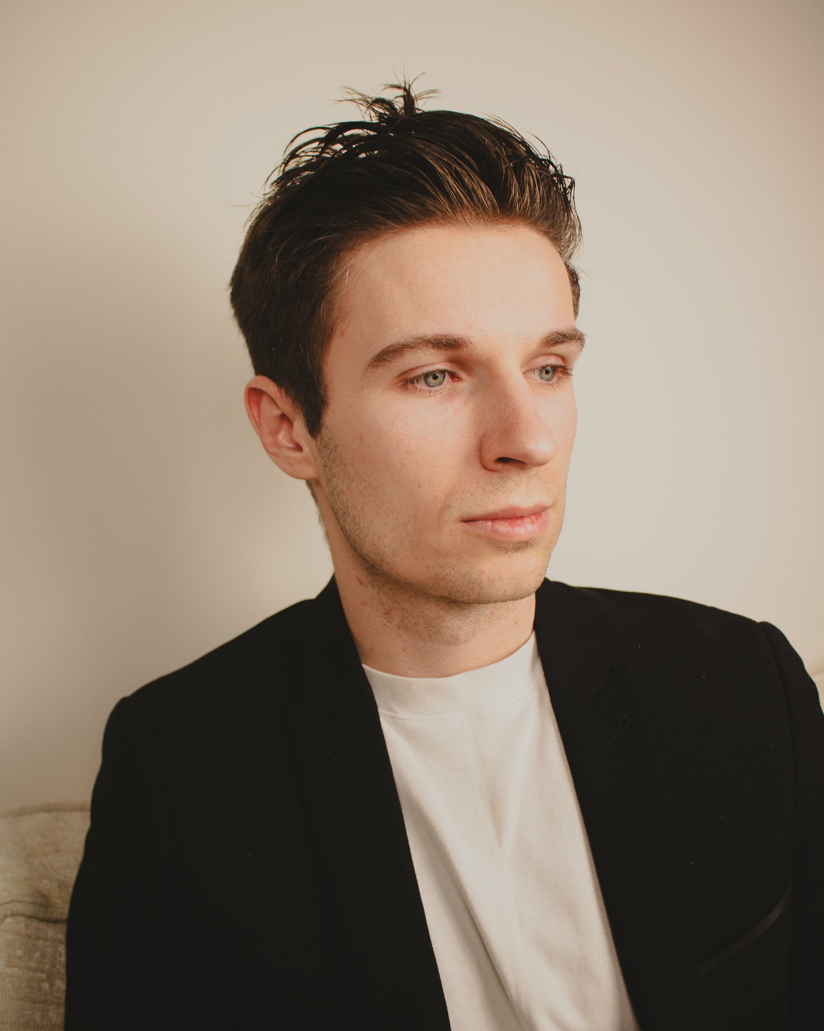 A young man with dark, styled hair and blue eyes wearing a black blazer over a white t-shirt, looking slightly to the side, against a plain light-colored background.