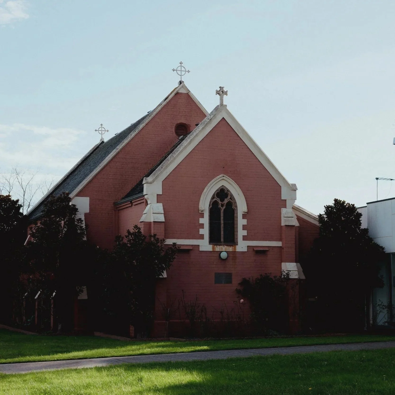 A pink brick church with pointed roof and crosses on top, set against a cloudy sky with trees and green grass in the foreground.