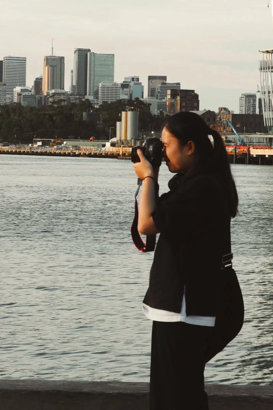 A woman with long black hair taking a photo with a camera near a body of water, with a city skyline of tall buildings in the background.