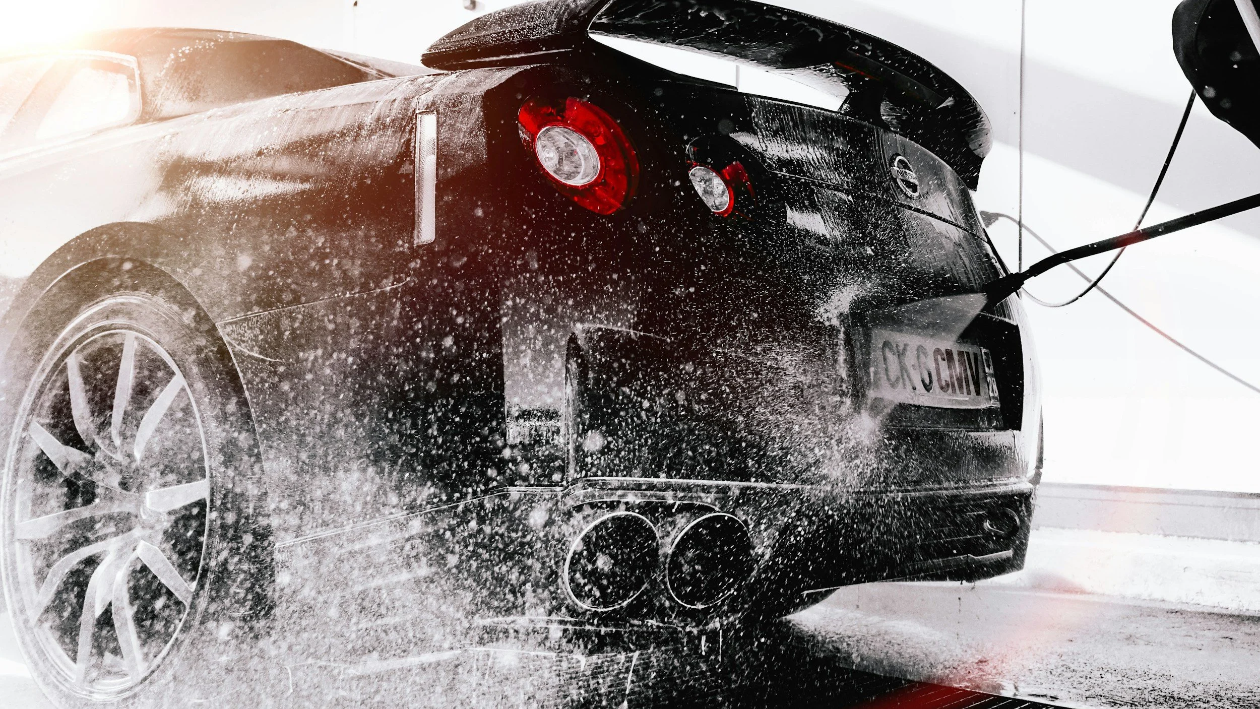 A black sports car being washed with soap and water, with soap suds on the surface and water splashing around. The car is indoors, and there is sunlight shining on the scene.