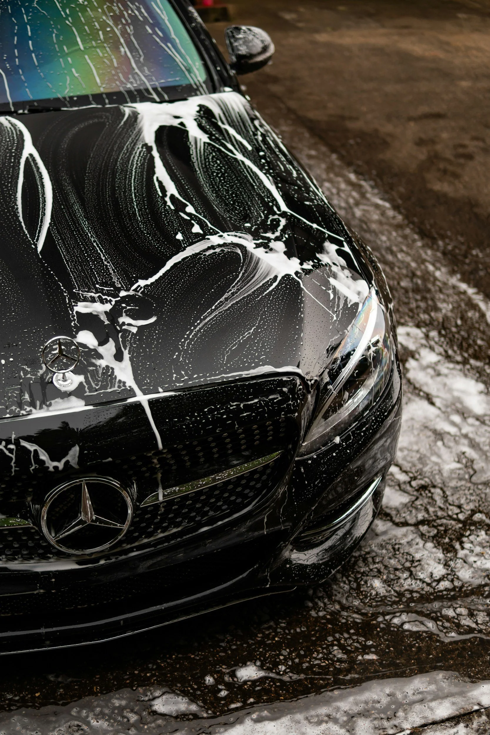 Front of a black Mercedes-Benz car covered with white soap suds, with soap dripping down the hood, on a wet, muddy ground.