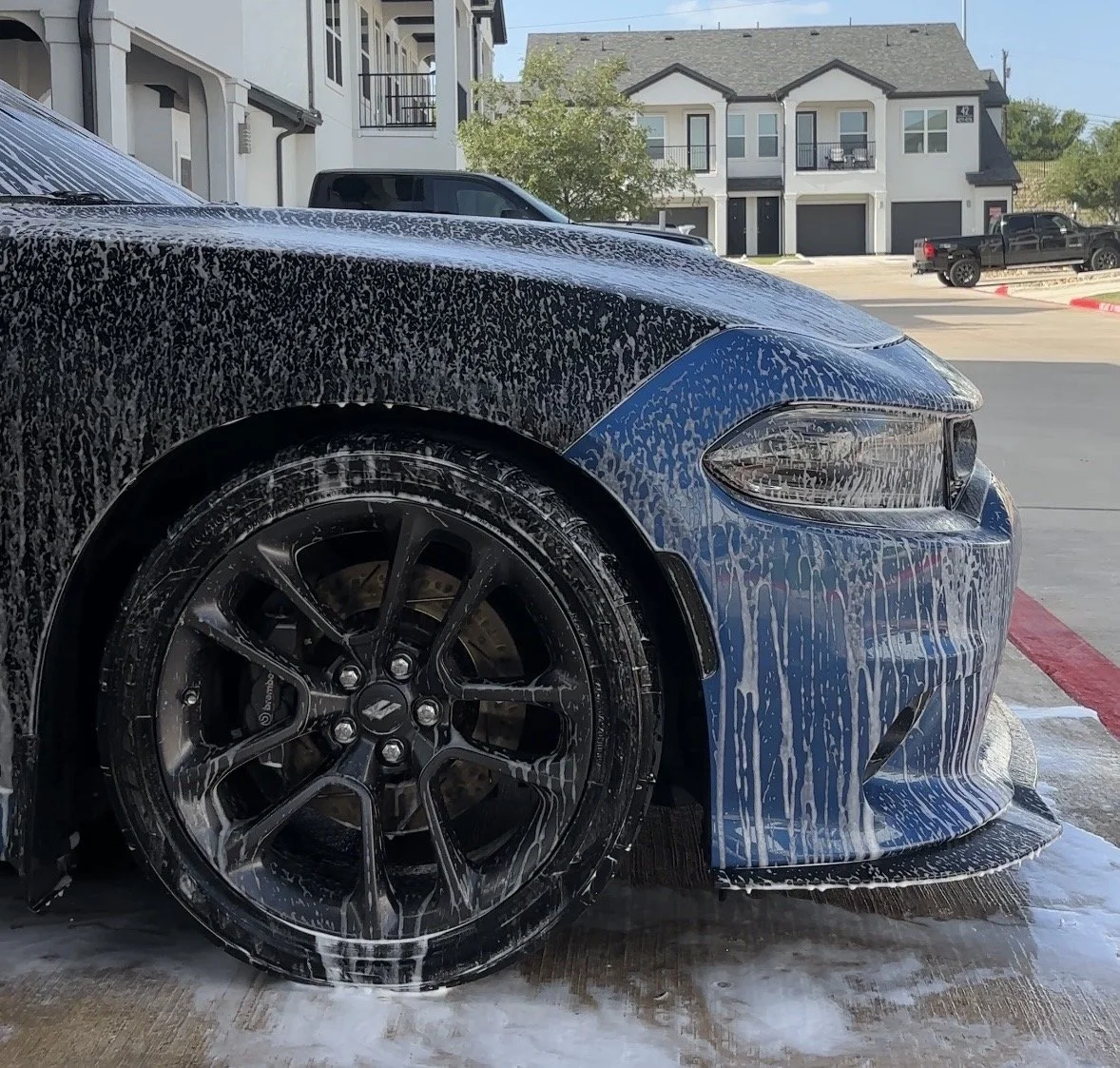 Close-up of a blue sports car being washed, with soap suds and water dripping down the front and the black wheel with spokes visible. In the background, apartment buildings and other vehicles are seen.