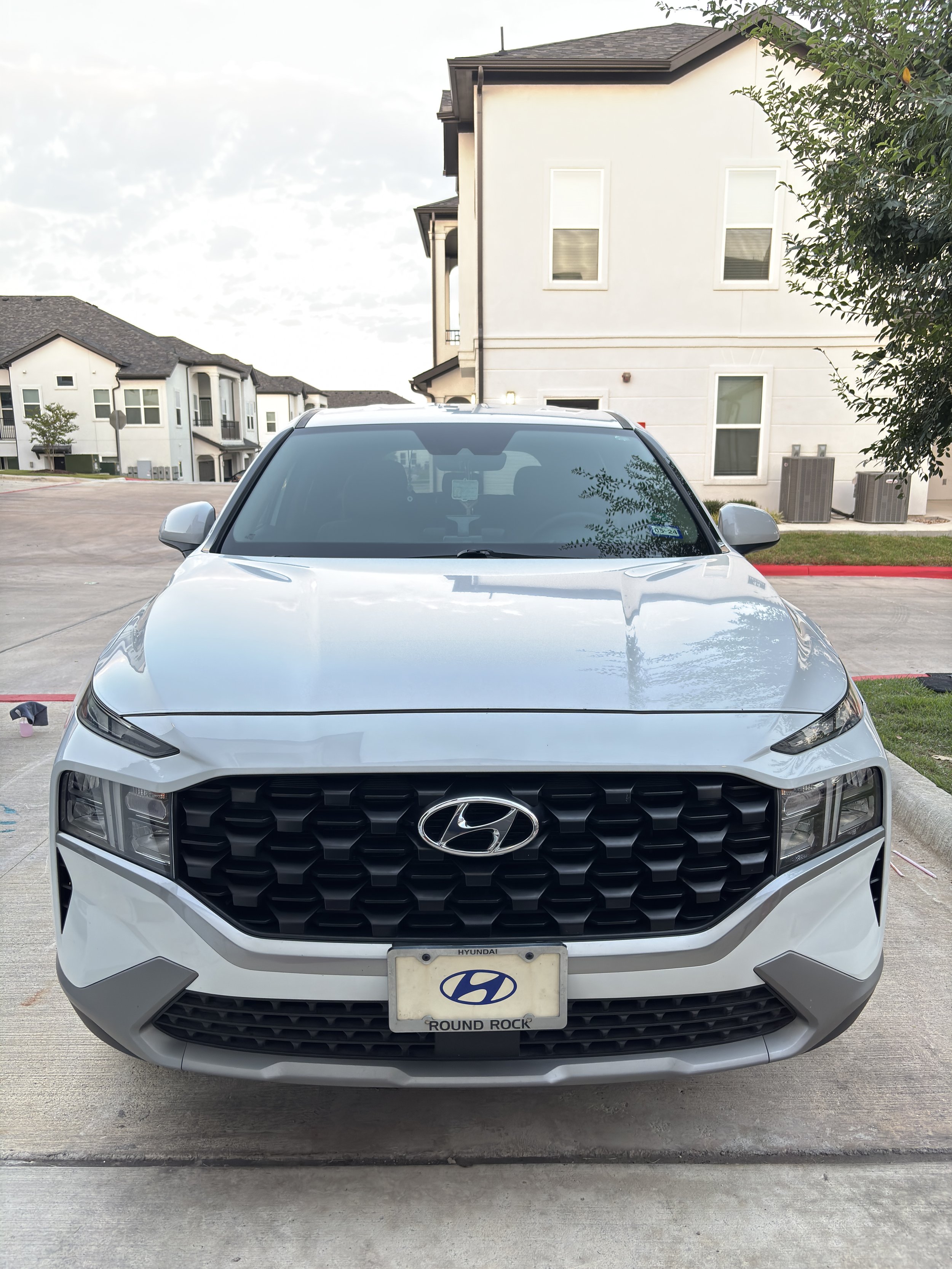 Front view of a white Hyundai SUV parked in a residential area with multi-story houses in the background and trees to the right.