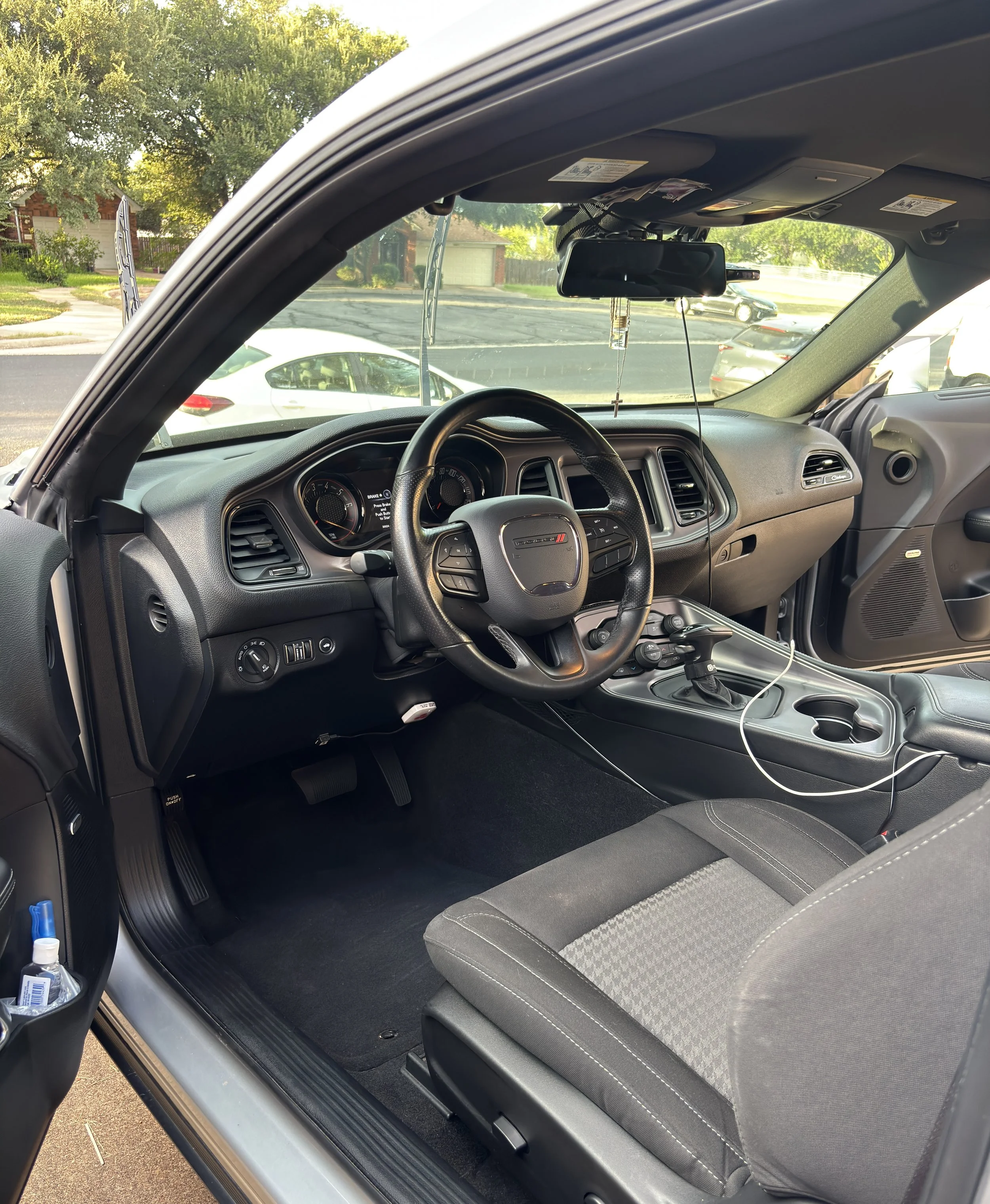 Interior view of a car with a black dashboard and a gray fabric seat. The steering wheel and gear shift are visible, along with a center console containing cup holders. A hanging accessory is attached to the rearview mirror. The front windshield shows trees and parked cars outside on a sunny day.