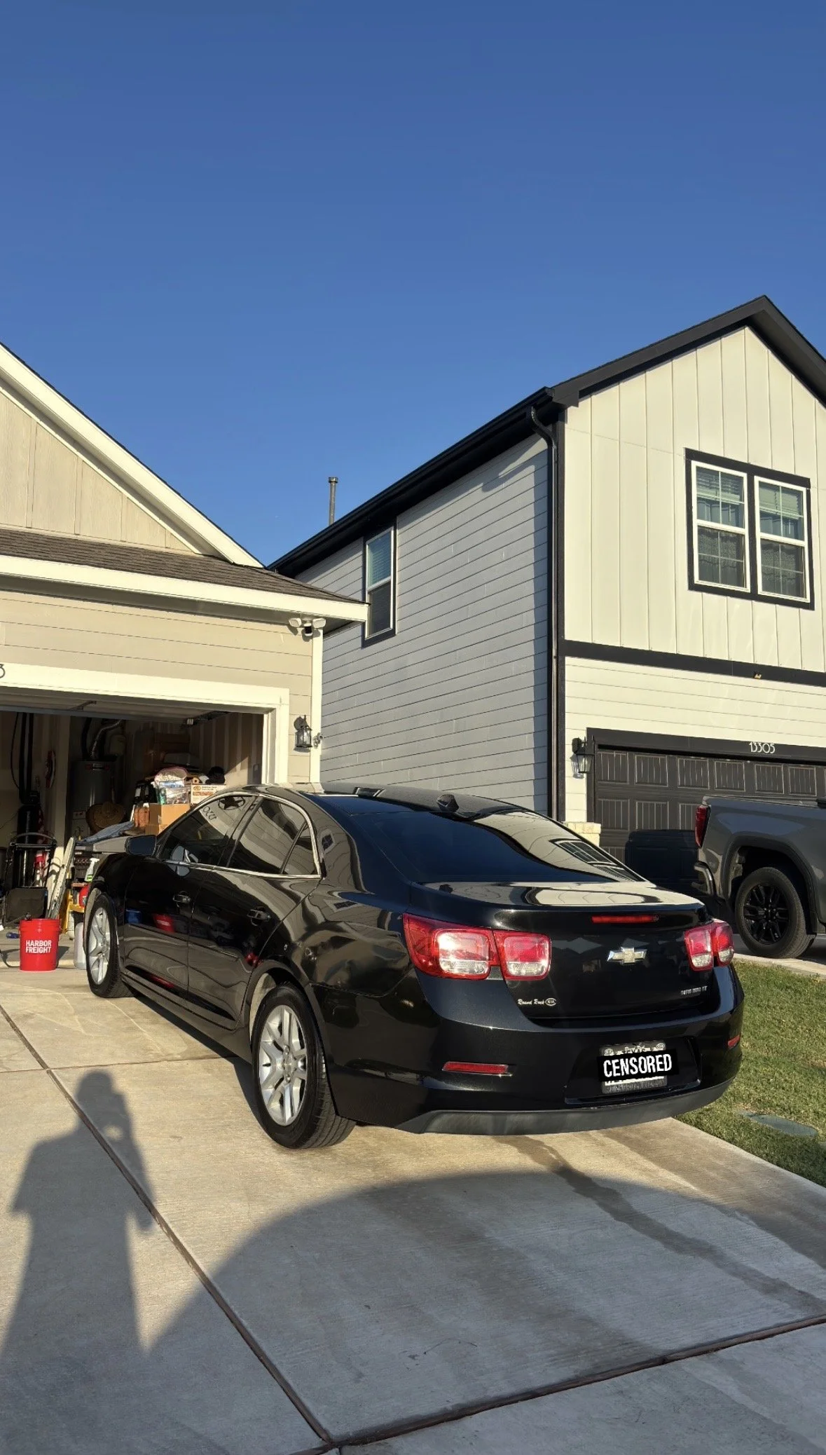 A black Chevrolet sedan parked in a driveway in front of a modern, two-story house with a garage, with a truck parked near the house. The houses have siding, and the sky is clear and blue.