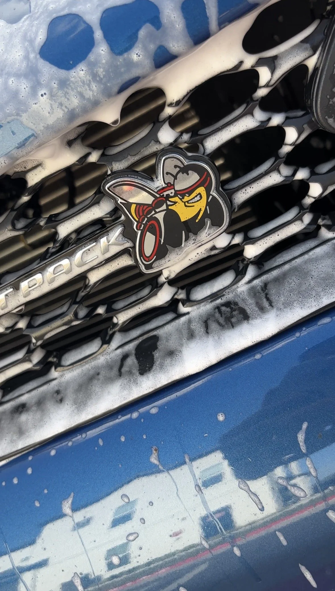 Close-up of a car front grille being washed with soap and water, with a cartoon bee sticker on the grille