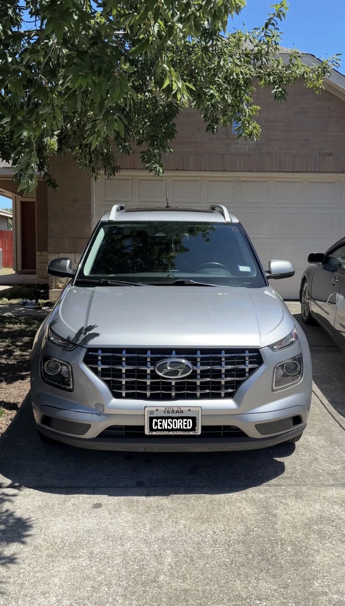 Silver Hyundai SUV parked in a driveway in front of a house with a brick exterior and garage door. A tree with green leaves partially shades the vehicle. The license plate is censored.
