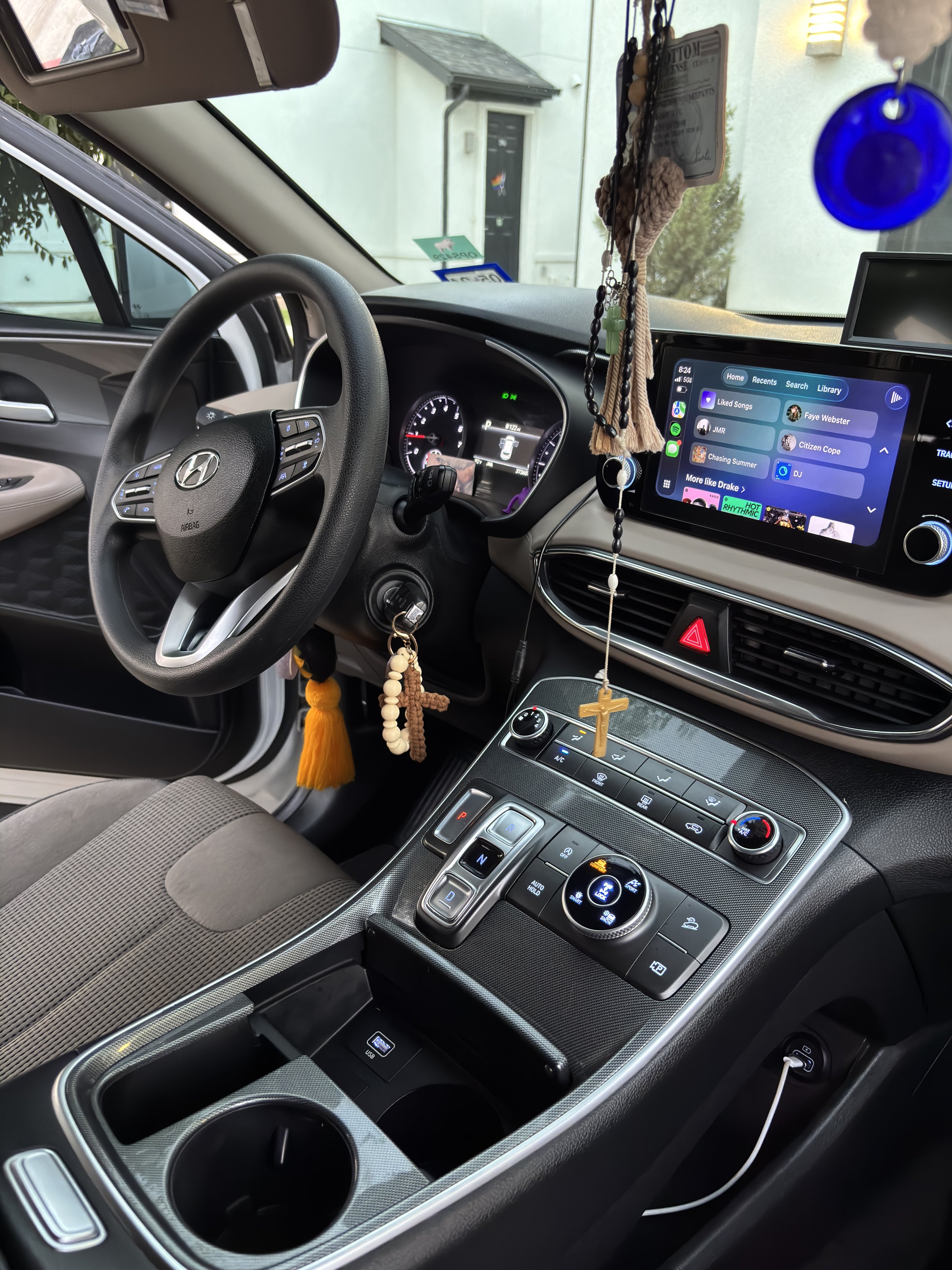 Interior of a Hyundai vehicle showing the steering wheel, digital dashboard, infotainment screen with music apps, hanging religious rosary and beads, gear shift, and various control buttons.