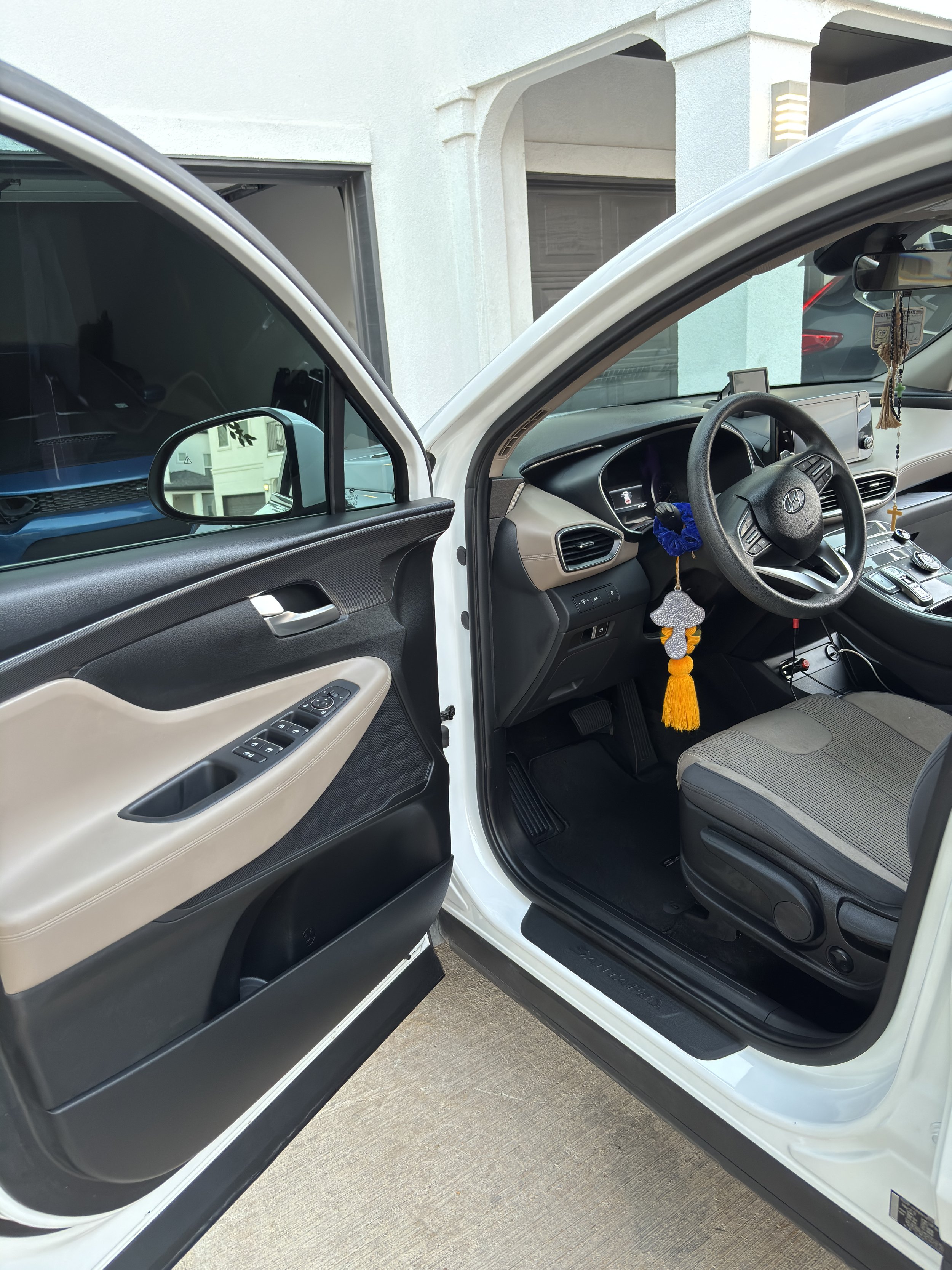 Open front door of a car showing gray interior with dashboard, steering wheel, and hanging decorations, parked on concrete driveway in front of a building with a garage door.