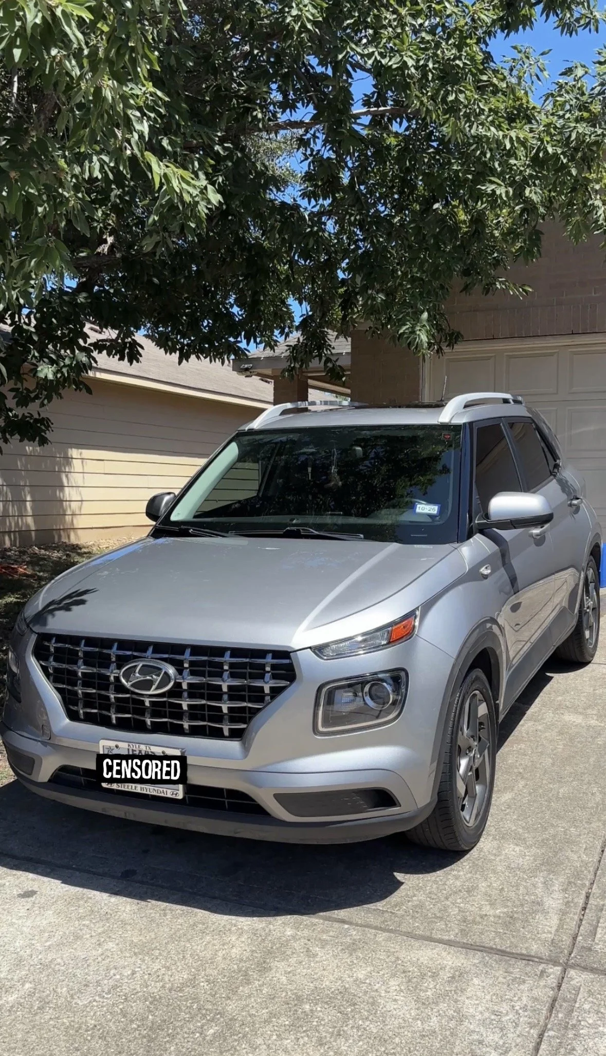 A silver Hyundai SUV parked in a driveway with a tree overhead and a house in the background.