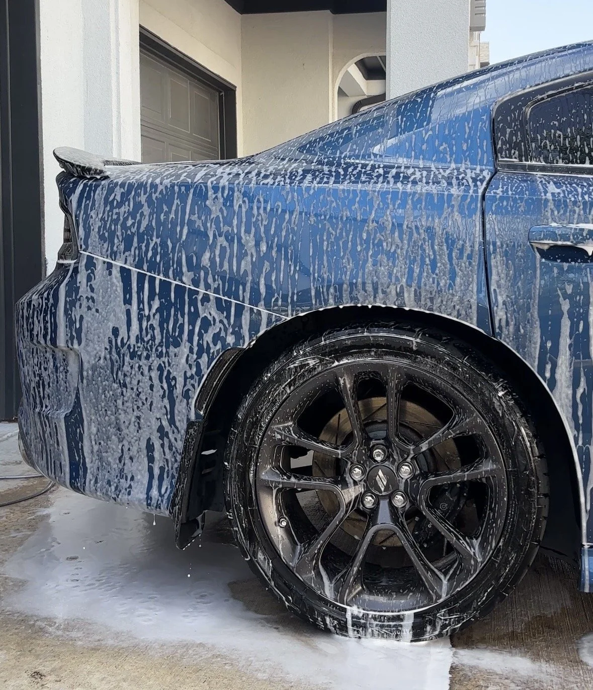 A blue sports car being washed with soap and foam, with soap suds covering the entire car and wheel.