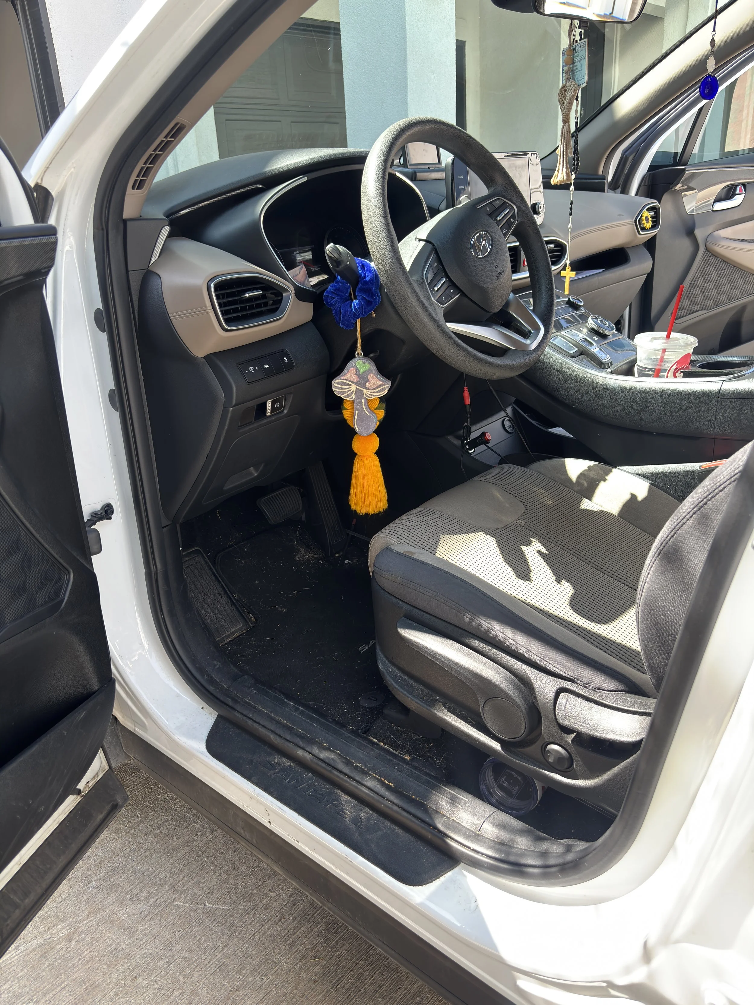 The interior of a white Hyundai vehicle showing the driver's seat, steering wheel, dashboard, and center console. The steering wheel has a blue plush scrunchie and hanging decor with a mushroom-shaped ornament and a yellow tassel. A drink cup with a red straw is placed on the console.