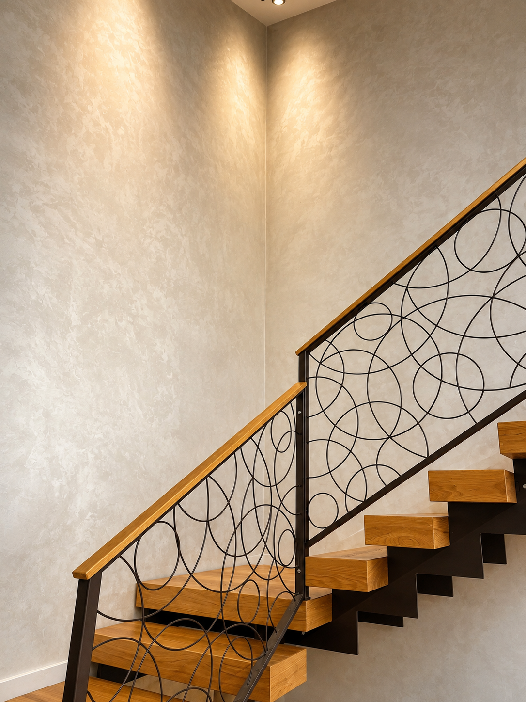 Scala in legno con ringhiera decorativa in metallo nero con cerchi concentrici, parete beige con finitura spazzolata, illuminazione soffusa.