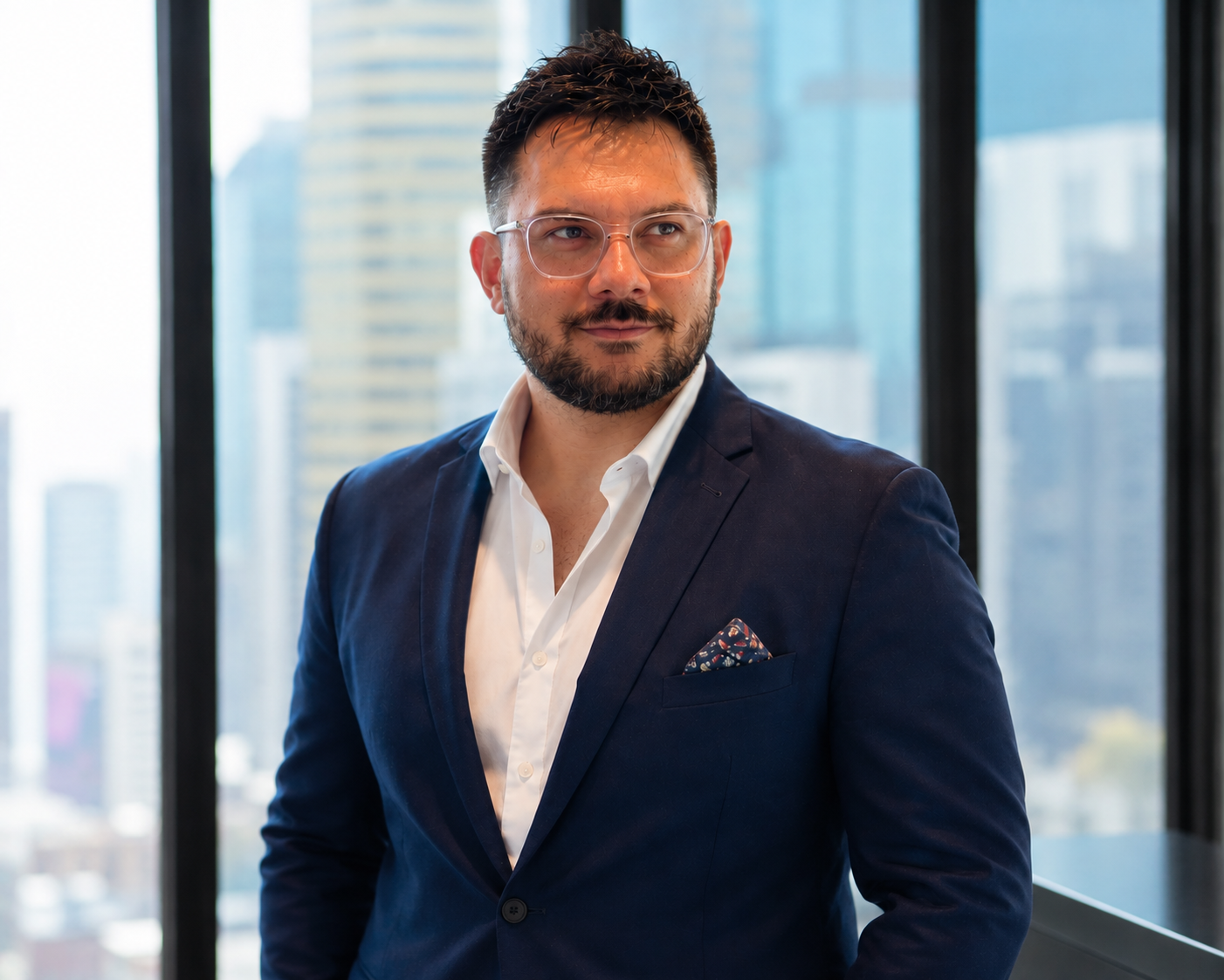 A man with glasses and a beard wearing a navy blue suit and white shirt, standing inside a modern office with city buildings visible through large windows in the background.