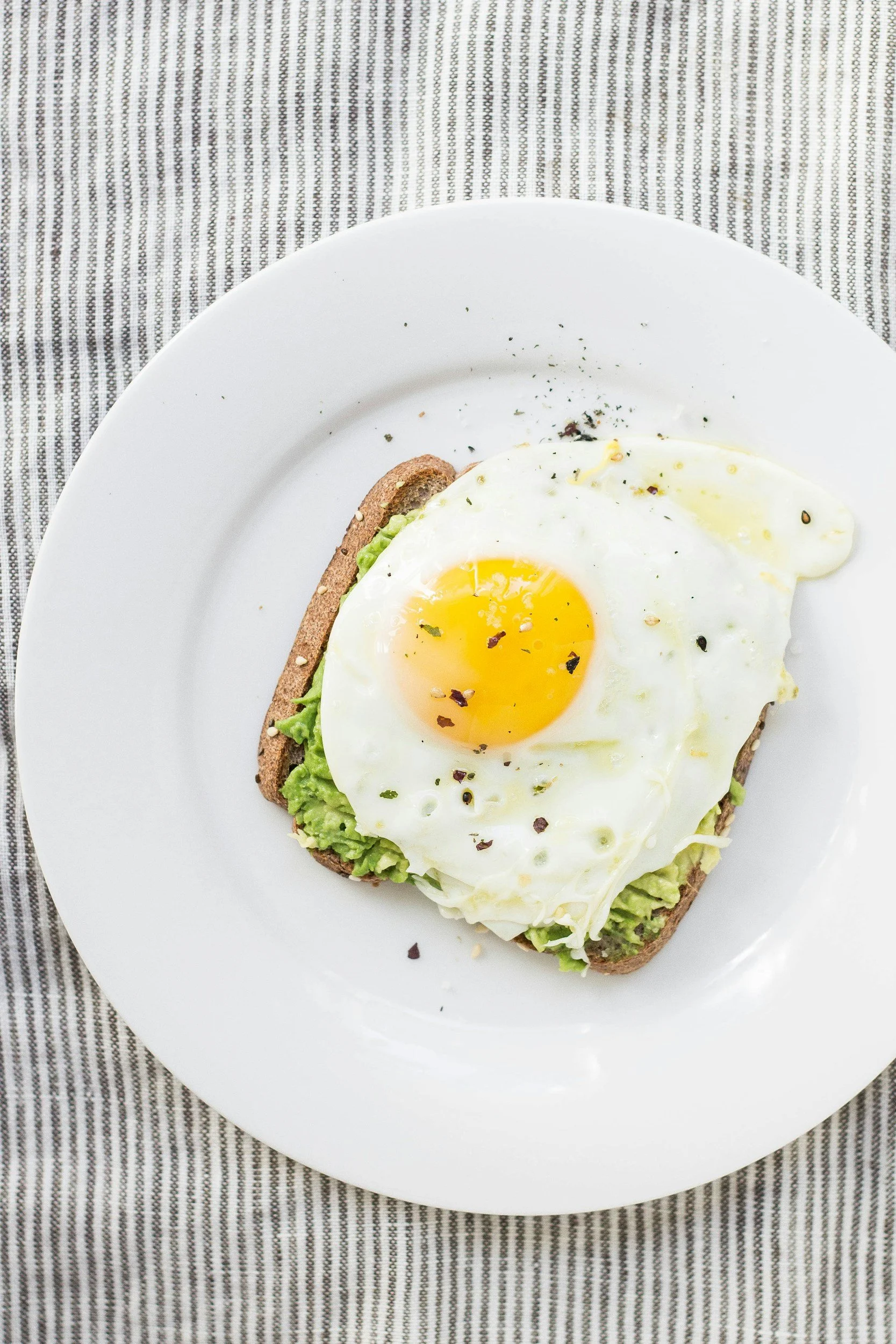 Whole grain toast topped with smashed avocado and a fried egg, sprinkled with spices, on a white plate resting on a gray striped tablecloth.