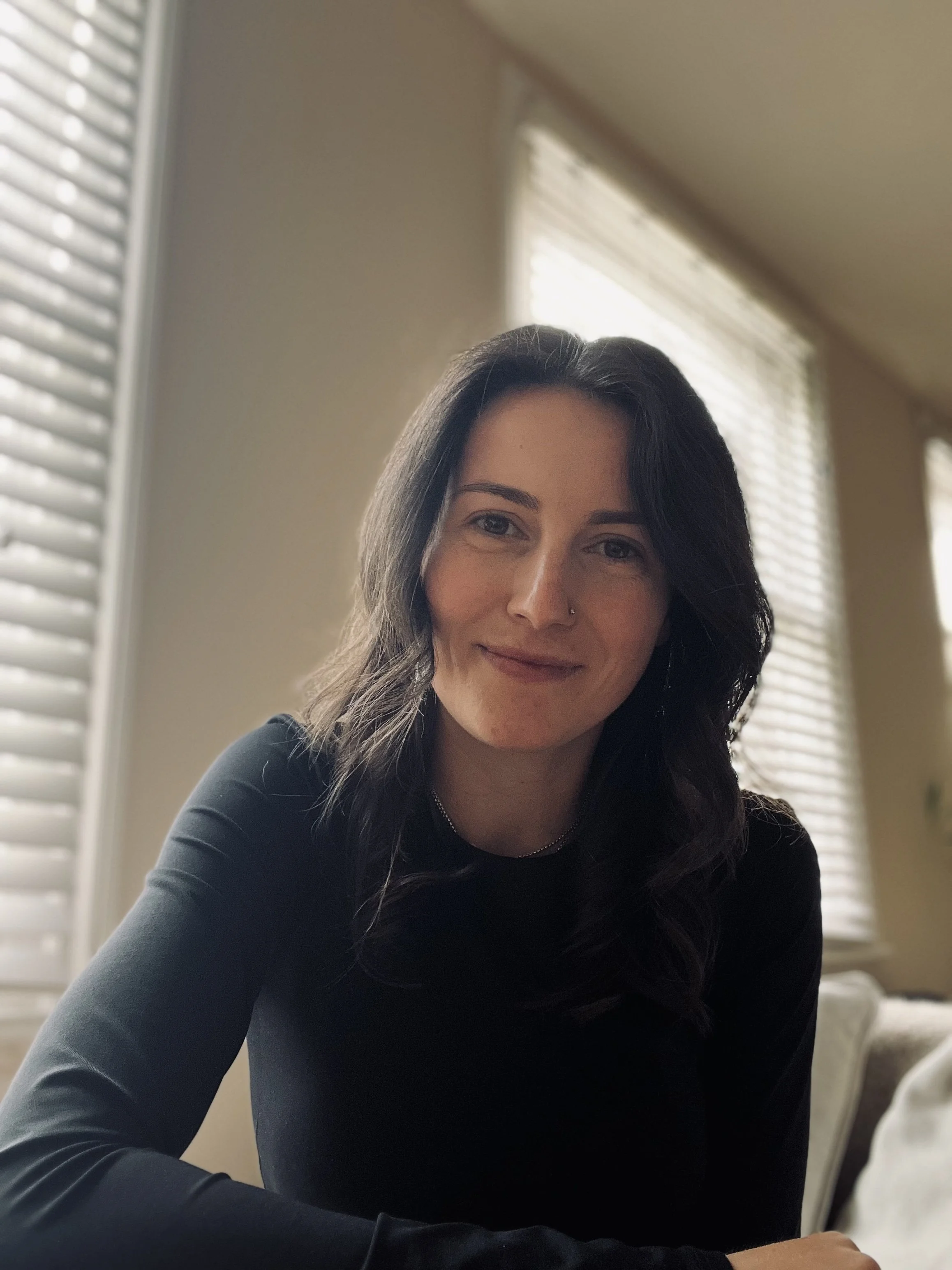 A woman with wavy dark hair and a nose piercing smiling in a room with beige walls and white blinds.