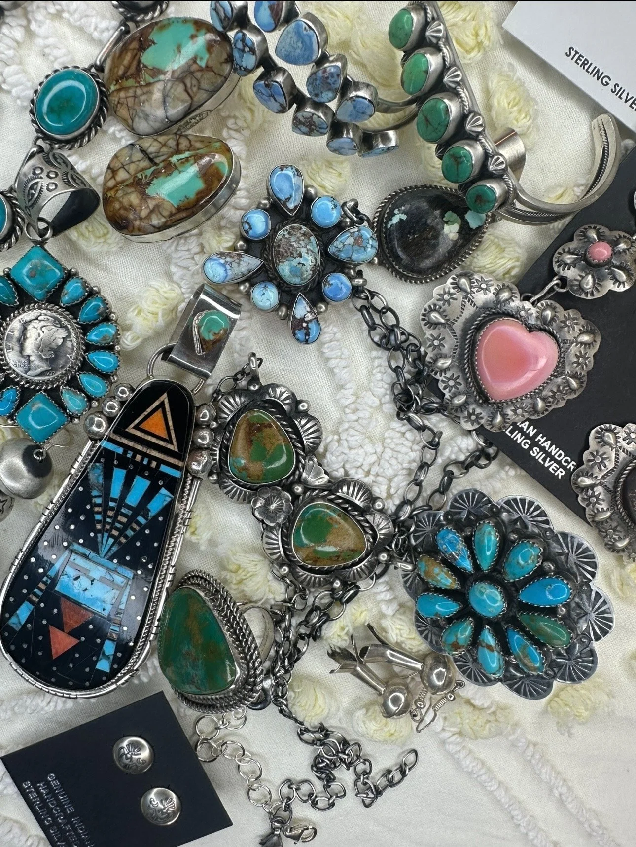 Collection of sterling silver jewelry, including turquoise and other stones, on a white fabric surface. The jewelry features various designs like rings, necklaces, and bracelets, some with intricate floral patterns and others with large oval or heart