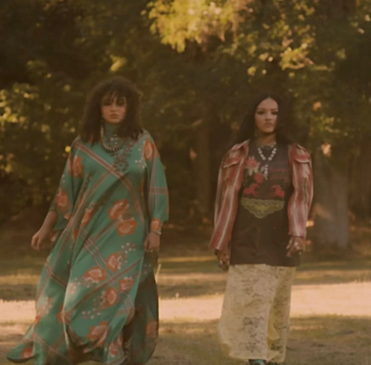 Two women walking outdoors in a park during sunset, wearing colorful bohemian-style outfits with jewelry, surrounded by trees with golden leaves.