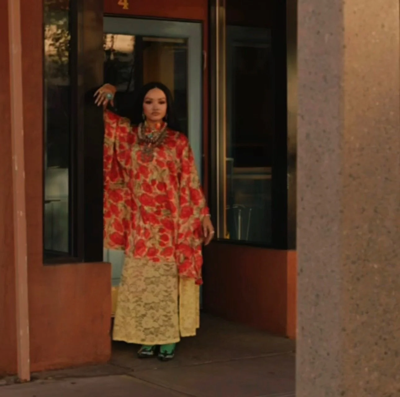 A woman wearing a vibrant red and gold traditional Asian dress, accessorized with jewelry, standing in a doorway.