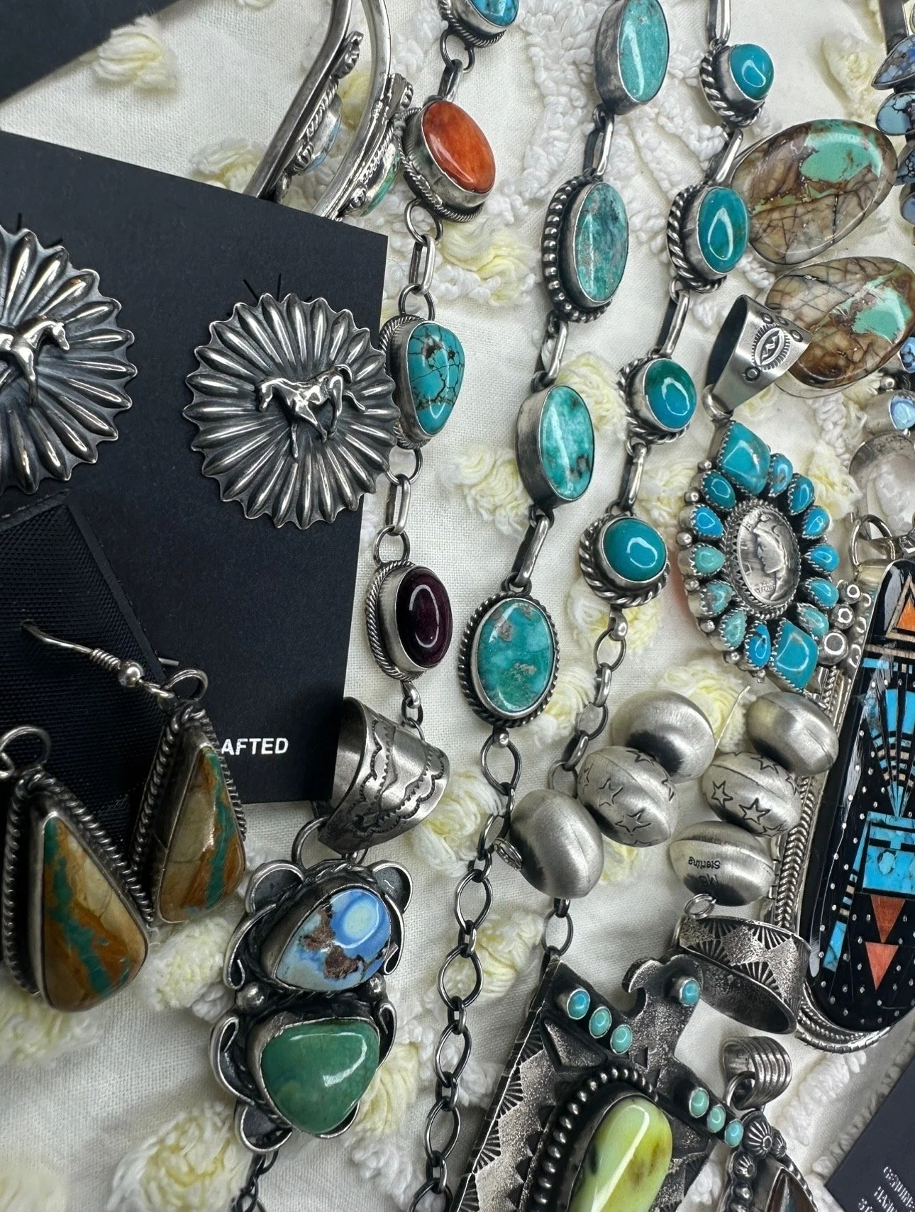 Various silver jewelry pieces with turquoise, coral, and other stones laid out on a white textured surface, including necklaces, bracelets, and earrings.