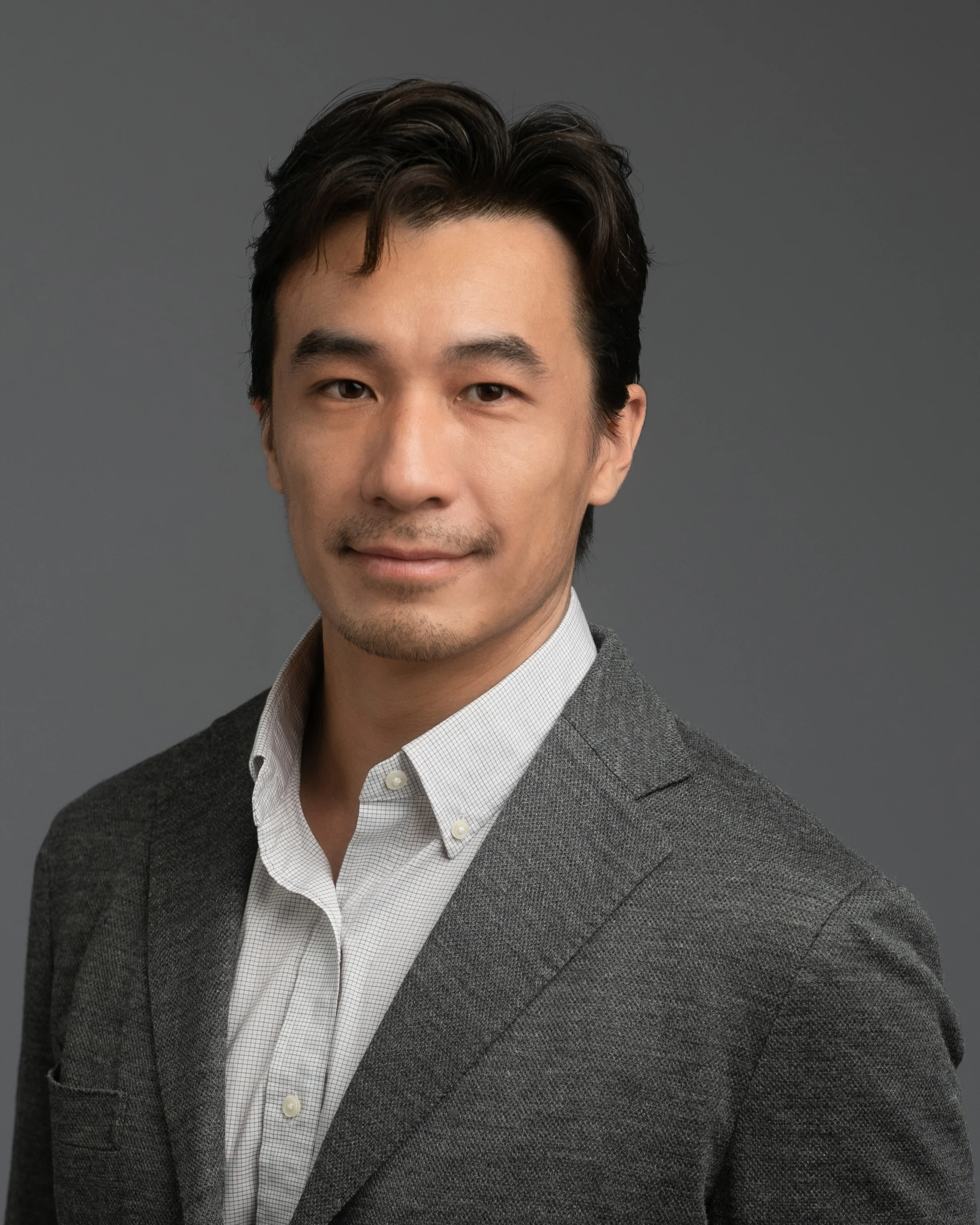 Close-up of a smiling Asian man wearing a gray blazer and white collared shirt against a gray background.