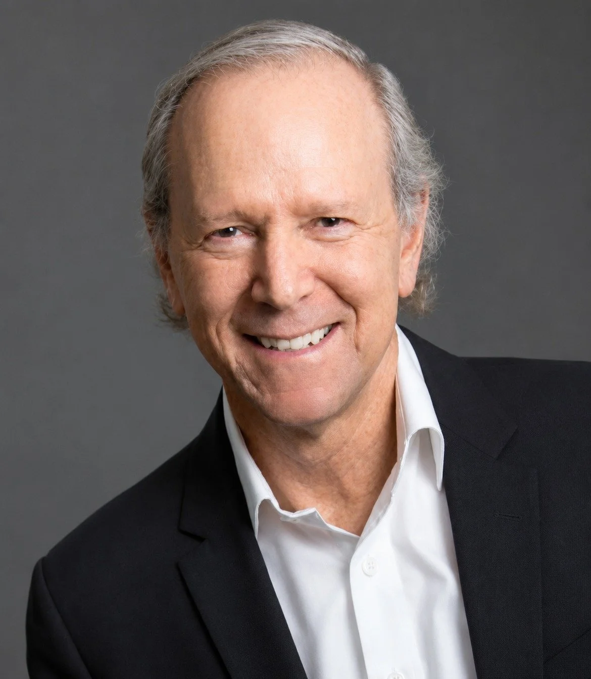 A middle-aged man with gray hair smiling, dressed in a black suit and white shirt, against a gray background.