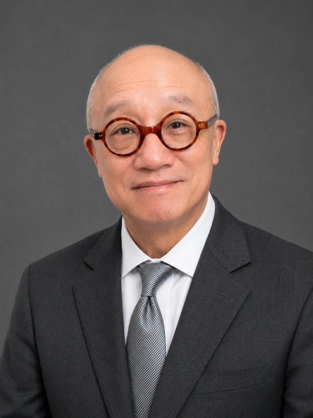 Headshot of an older Asian man with glasses, dressed in a dark suit, white shirt, and patterned tie, against a gray background.