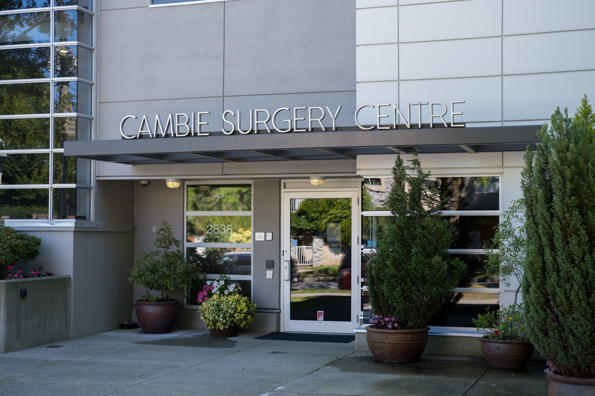 The entrance of a modern medical building with signs reading 'Cambie Surgery Centre.' There are potted plants and flowers outside, with a glass door and large window on the facade.