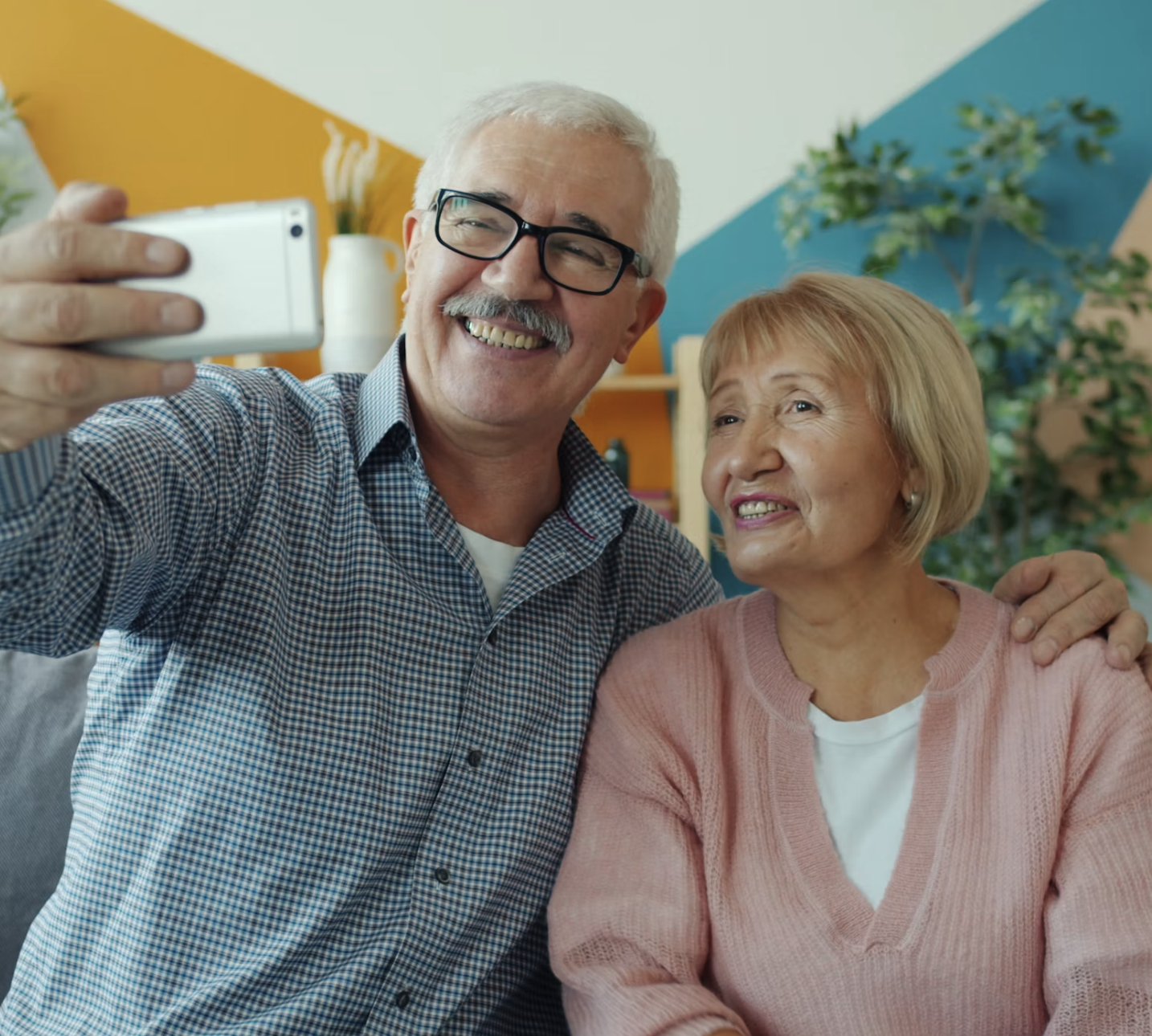 An elderly man with gray hair, glasses, and a checkered shirt is taking a selfie with a smiling elderly woman with blond hair, wearing a pink sweater. They are indoors with colorful walls and plants in the background.
