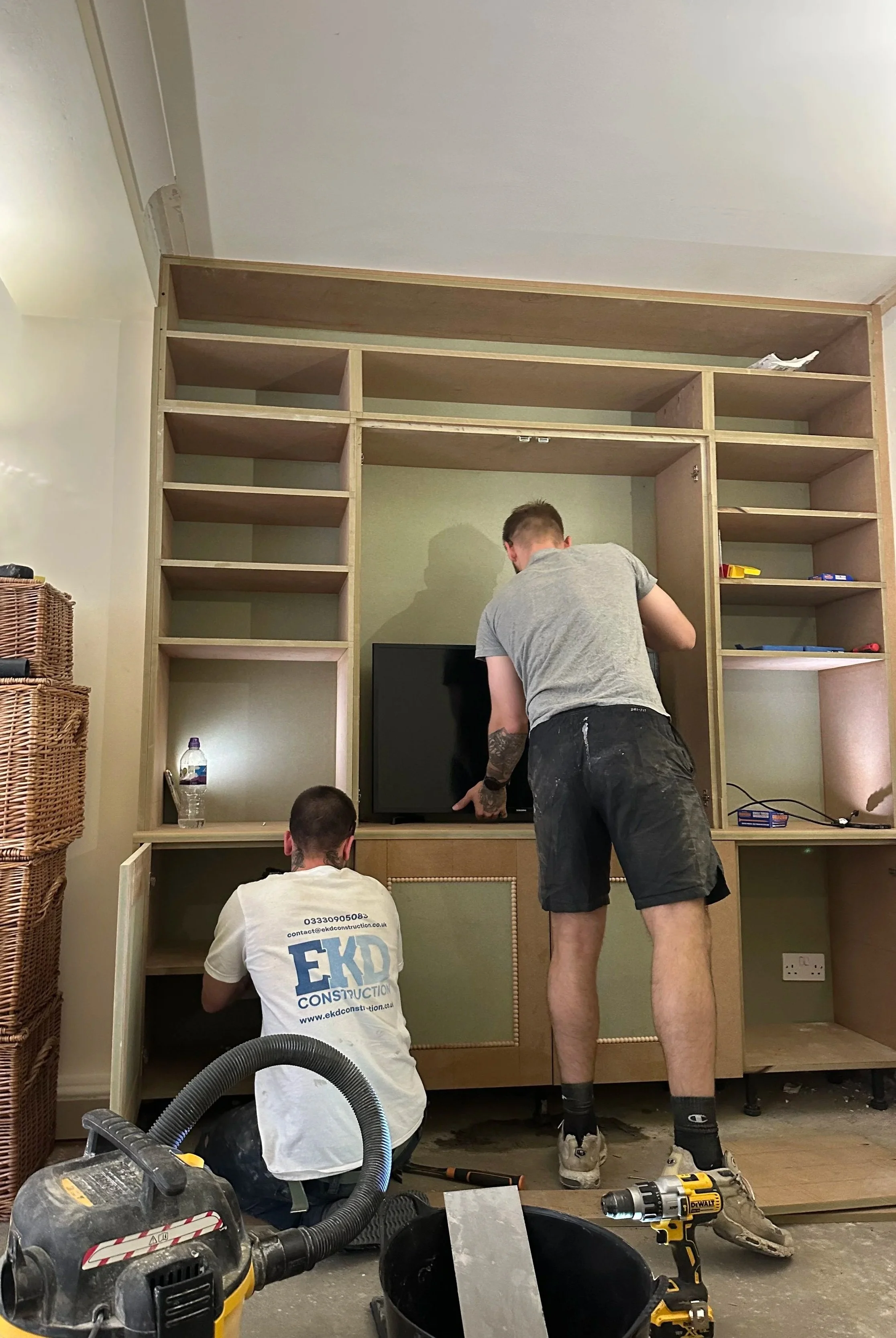 Two men are building a wooden entertainment center with open shelves and cabinets. One man is kneeling, working inside the lower cabinet, while the other is standing and adjusting the top part of the construction. Tools and materials, including a cordless drill, vacuum, and screws, are scattered around them.