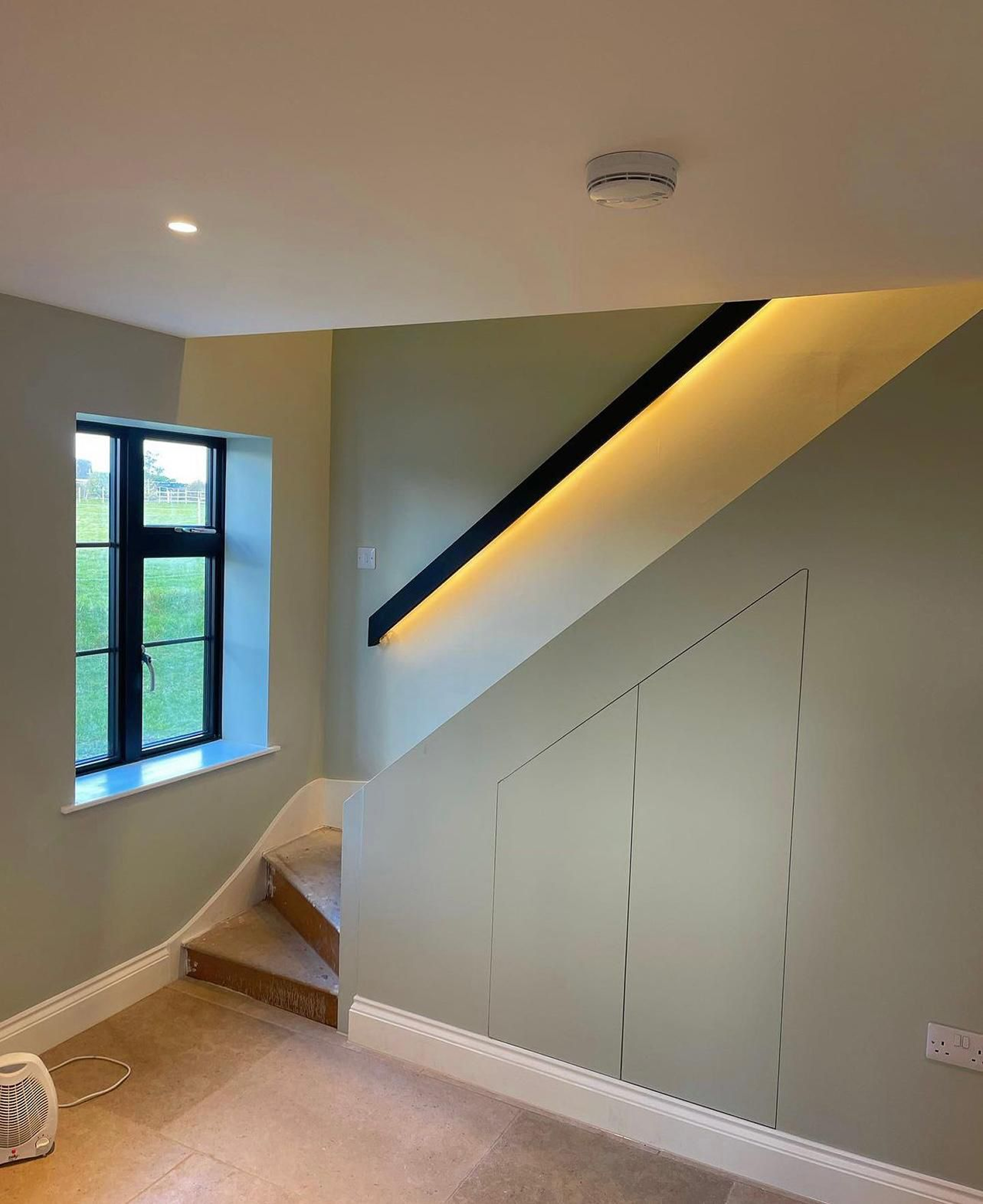 Interior view of a room with a window showing a greenery outside, a staircase with wooden steps, a built-in cabinet painted light green with a black diagonal trim with yellow lighting underneath, a smoke detector on the ceiling, and electrical outlets on the wall.