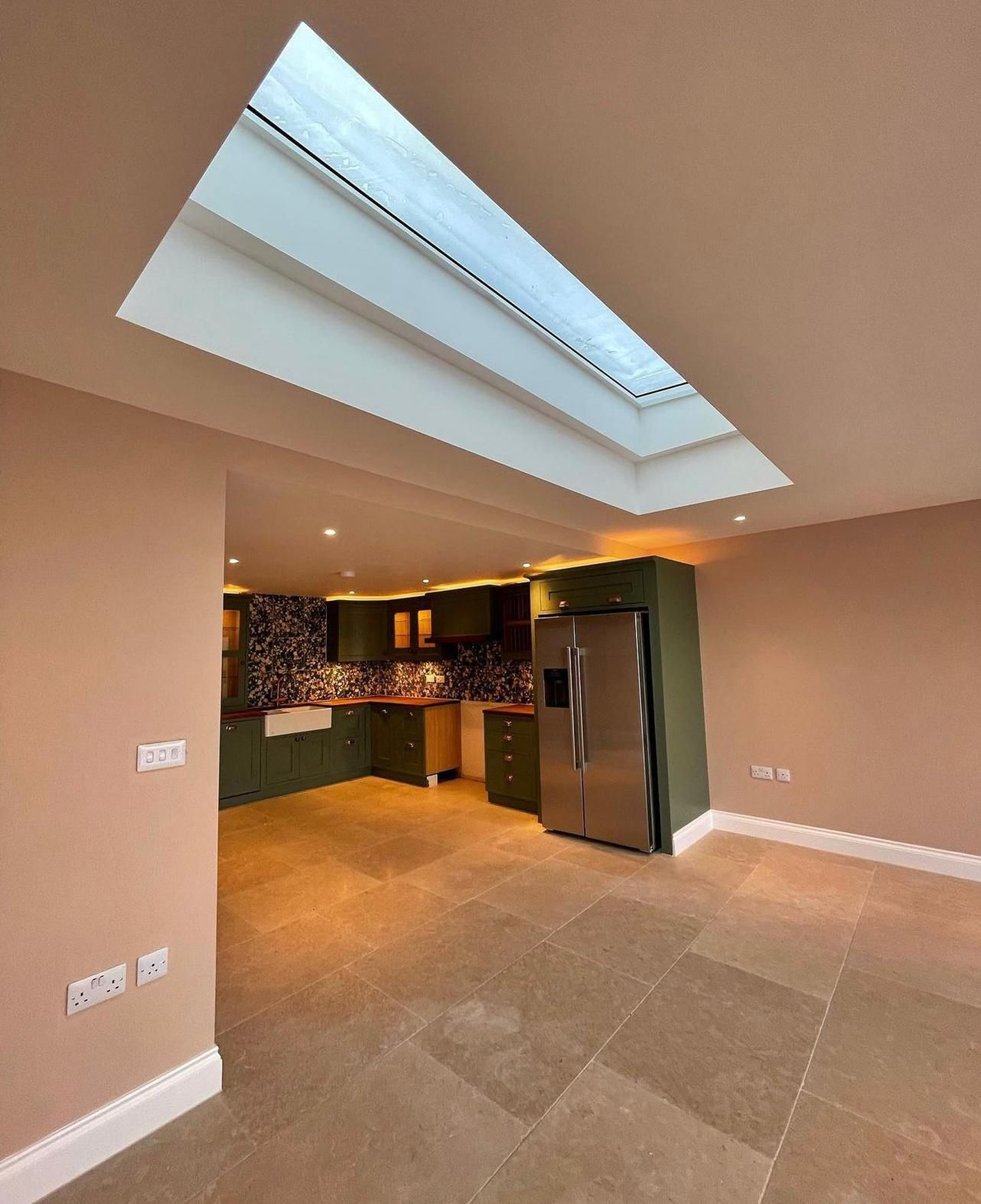 Interior view of a kitchen with beige walls, green cabinets, stainless steel refrigerator, tiled floor, and a skylight ceiling.