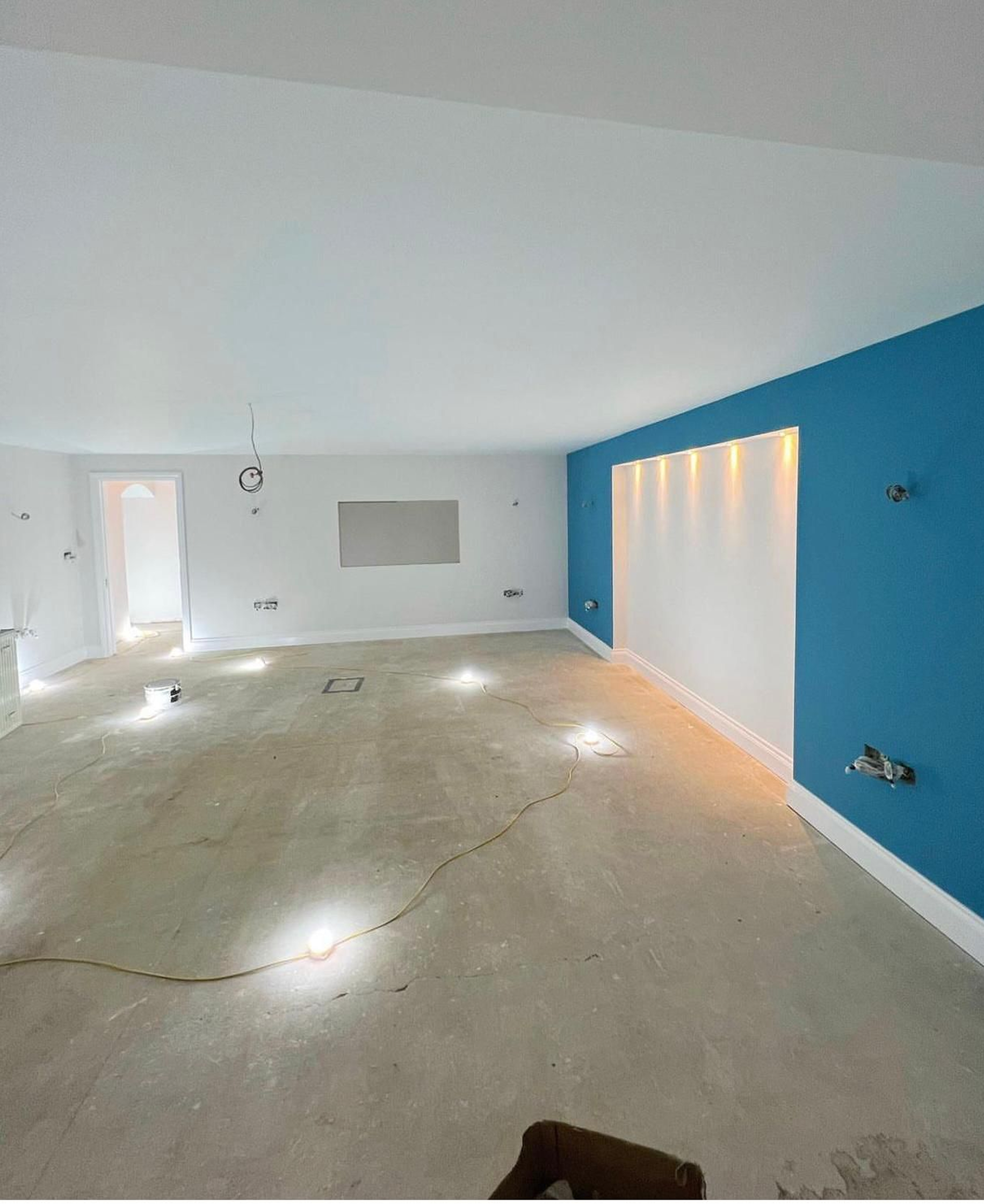 A room with a skylight on the ceiling, a blue accent wall with built-in lighting, hardwood flooring, and some construction tools and lighting equipment on the floor.