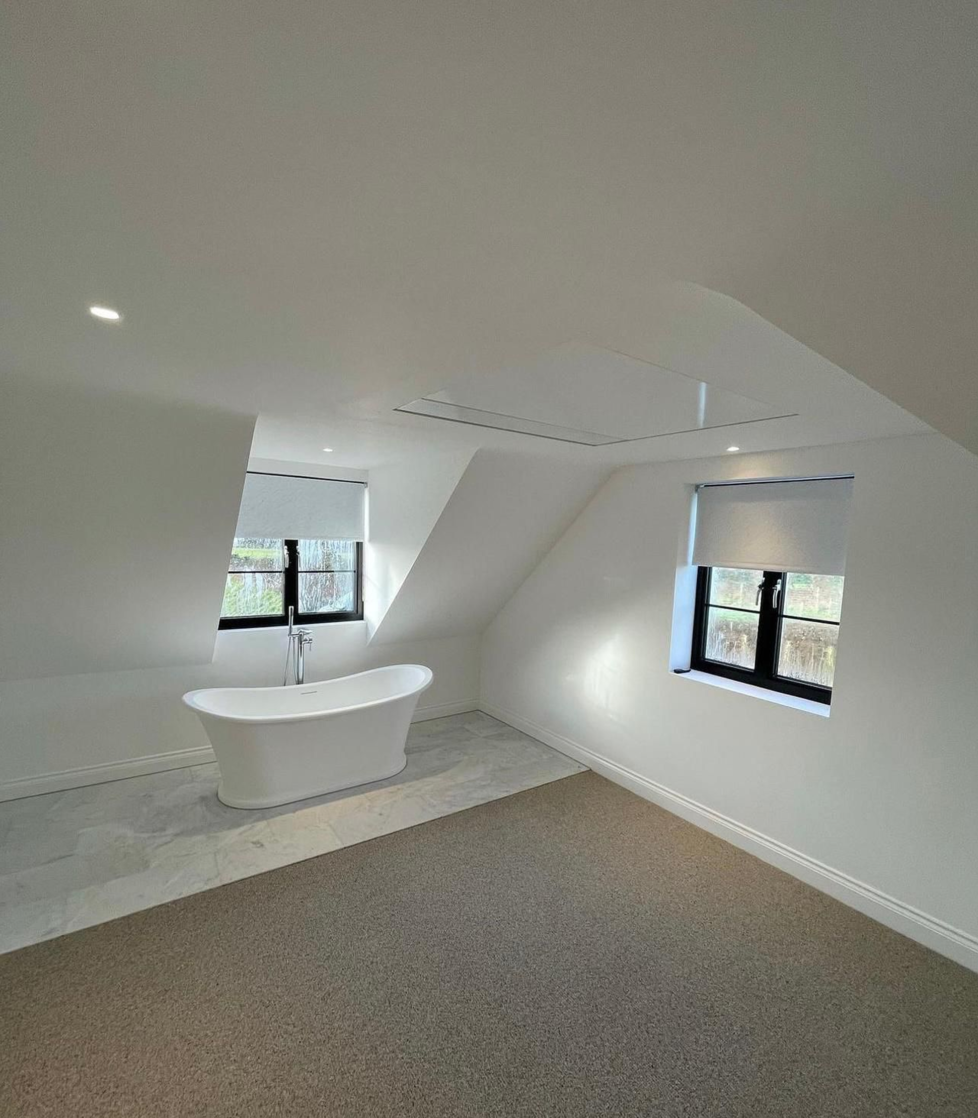 Modern bathroom with a freestanding white bathtub on a marble platform, dark window frames, and sloped ceiling with built-in lighting.