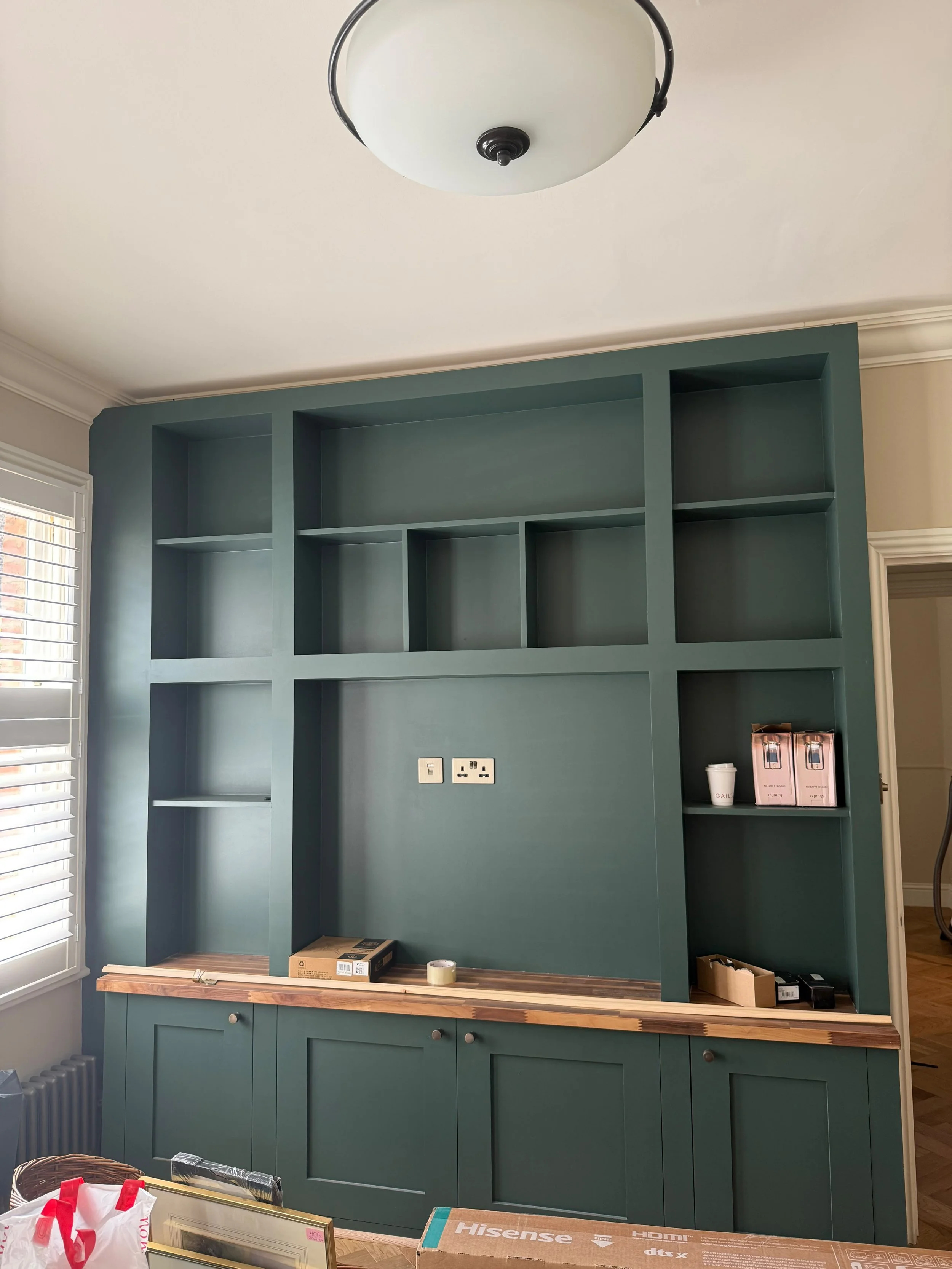 An empty teal-blue built-in bookshelf with open shelves and a wooden countertop below, in a room with beige walls and a window with white blinds. Items on the countertop include boxes, a roll of tape, and a small cup.