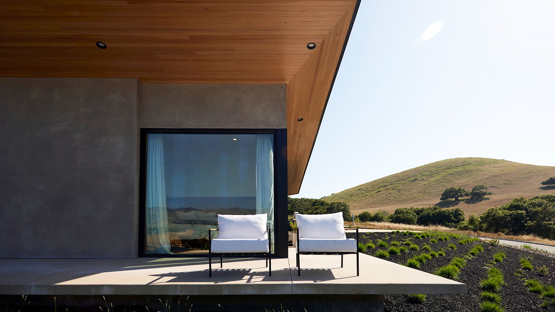 Modern house exterior with large window and outdoor seating, hillside landscape in the background.