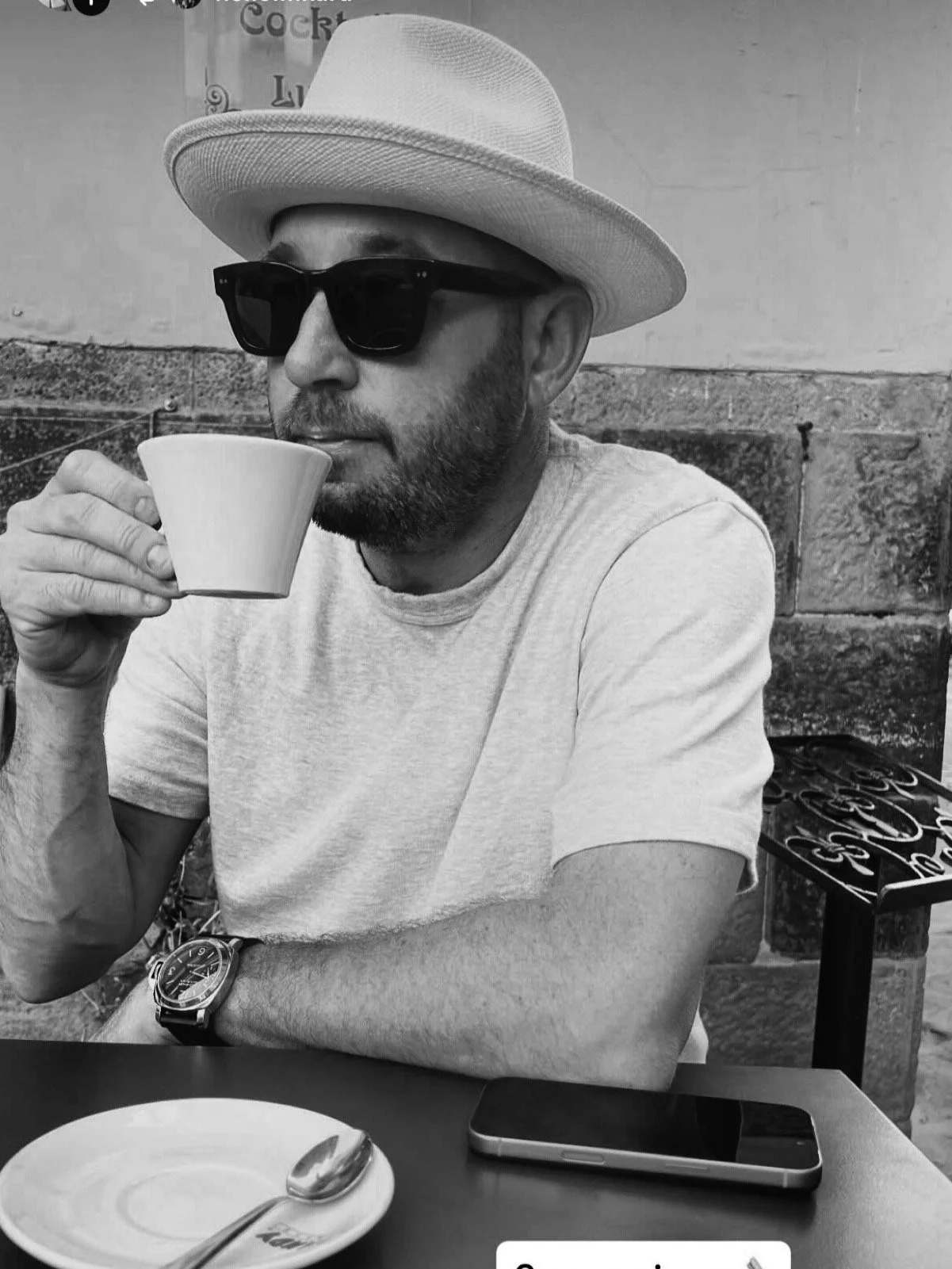 A man wearing sunglasses and a hat, holding a coffee cup near his mouth, sitting at a table with a plate, spoon, and a smartphone in front of him, outdoors with a stone wall background.