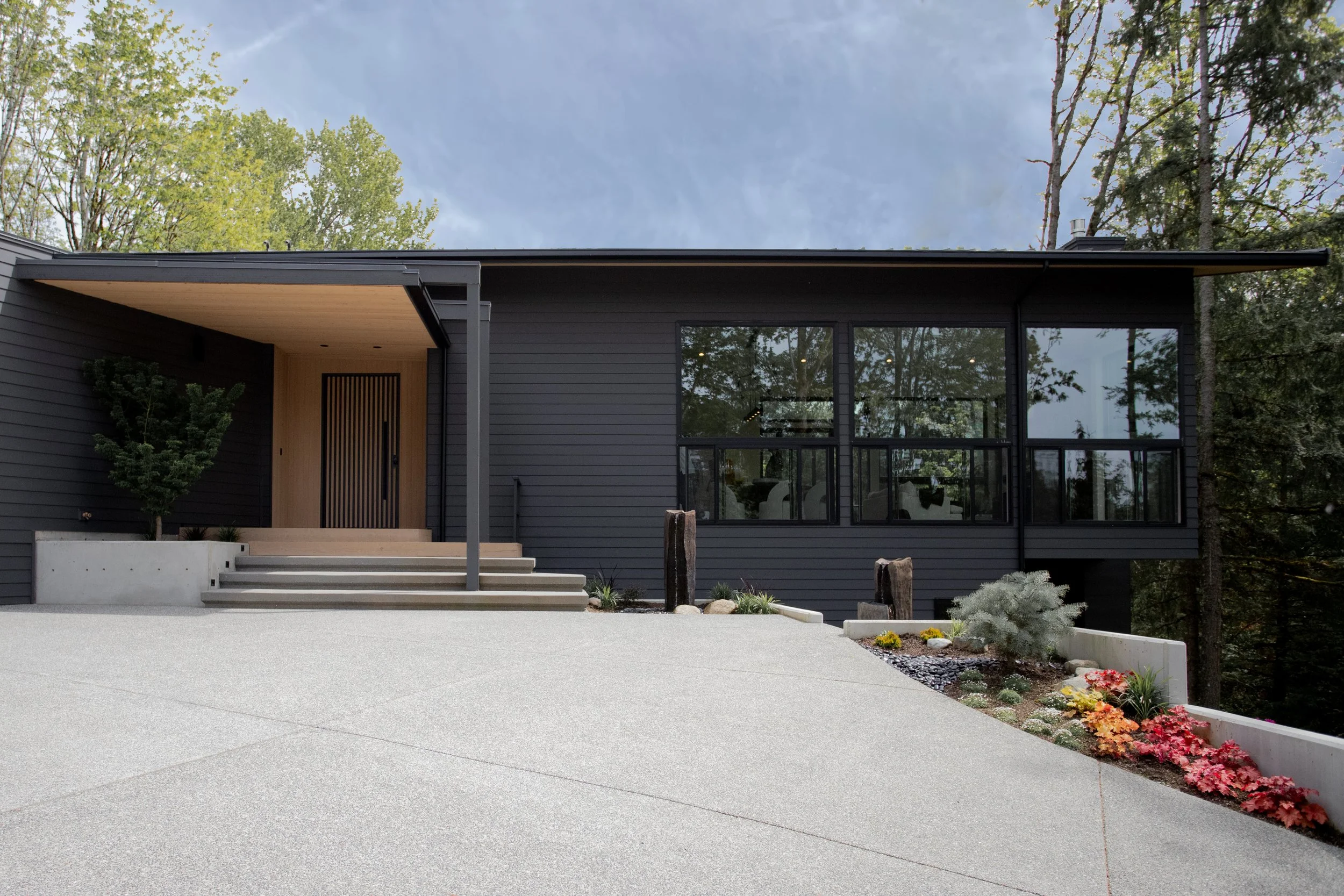 Modern black house with large windows, a front porch, concrete steps, landscaped garden with colorful flowers and small shrubs, surrounded by trees under a cloudy sky.