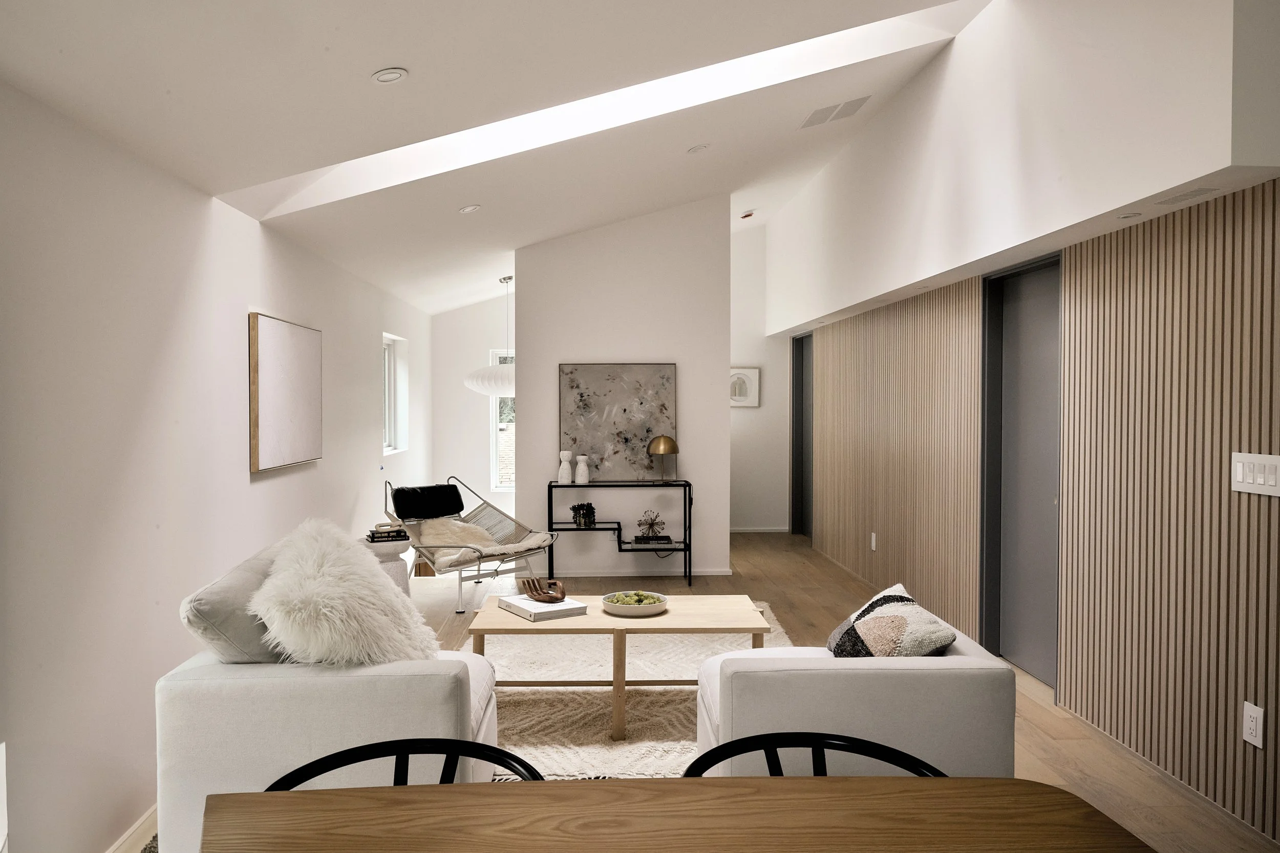 Modern living room with white walls, natural wood accents, and minimalistic decor. Seating includes two cream armchairs and a sofa with throw pillows. There is a light wooden coffee table with decorative items, and a black bench with white fur throw.