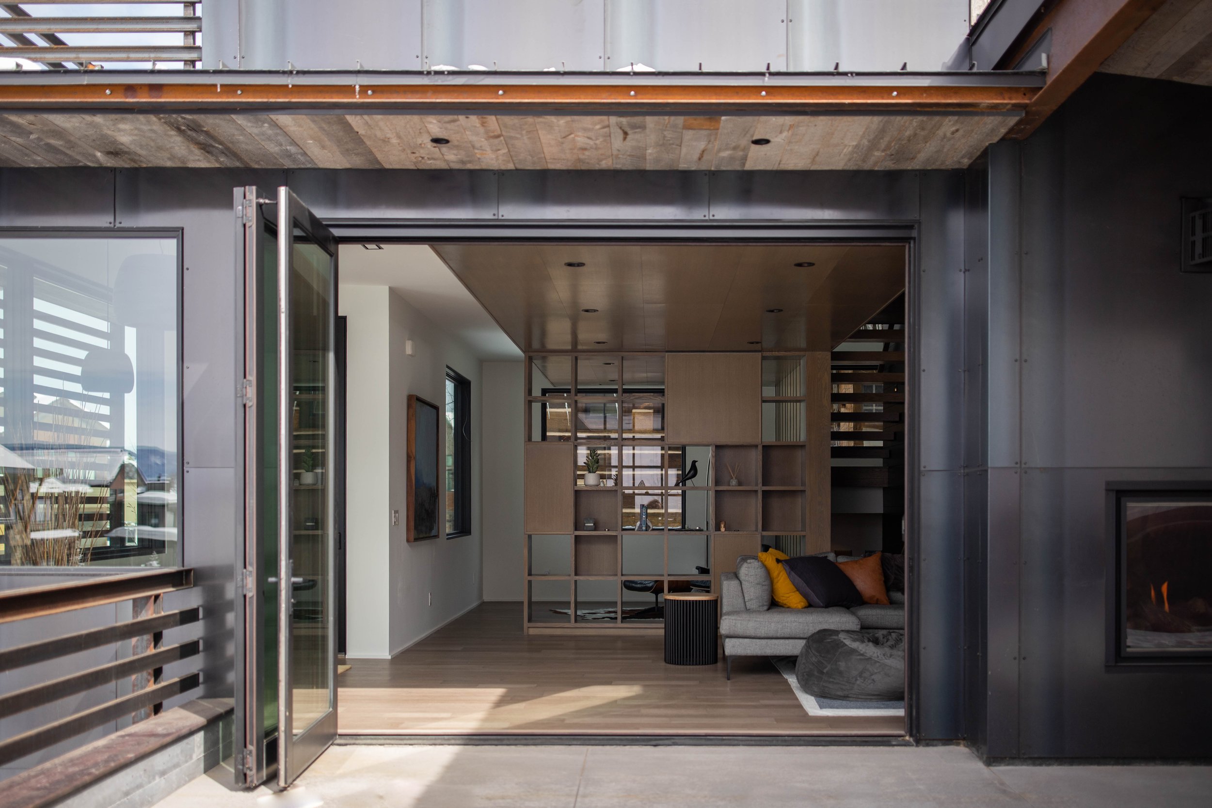 Modern living room with open sliding glass doors, wooden ceiling, and a mix of contemporary furniture including a sofa with yellow and orange cushions, a black side table, and a wooden shelving unit with decor items.