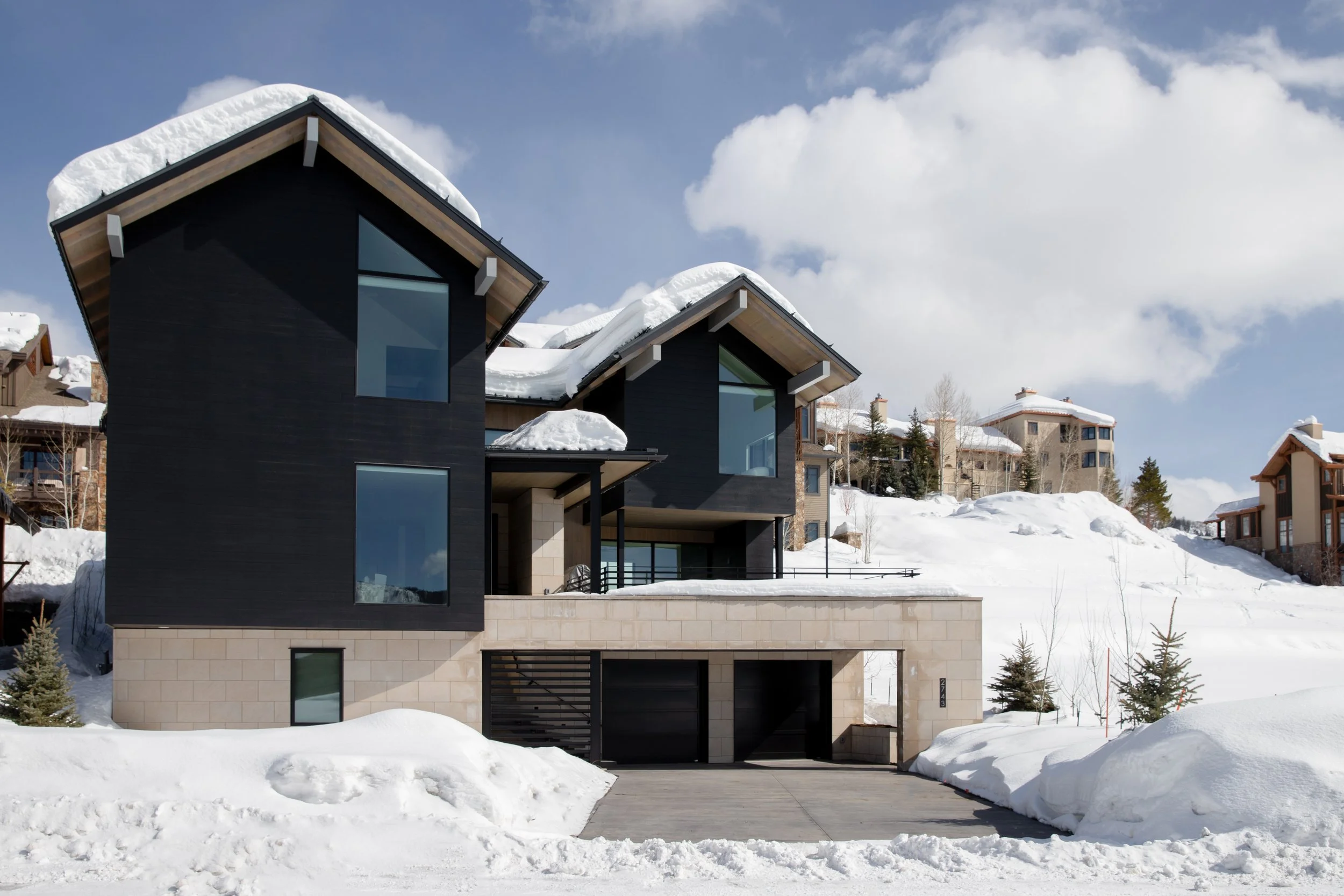 Modern black and beige house with large windows and a snow-covered roof, set in a snowy landscape under a partly cloudy sky.