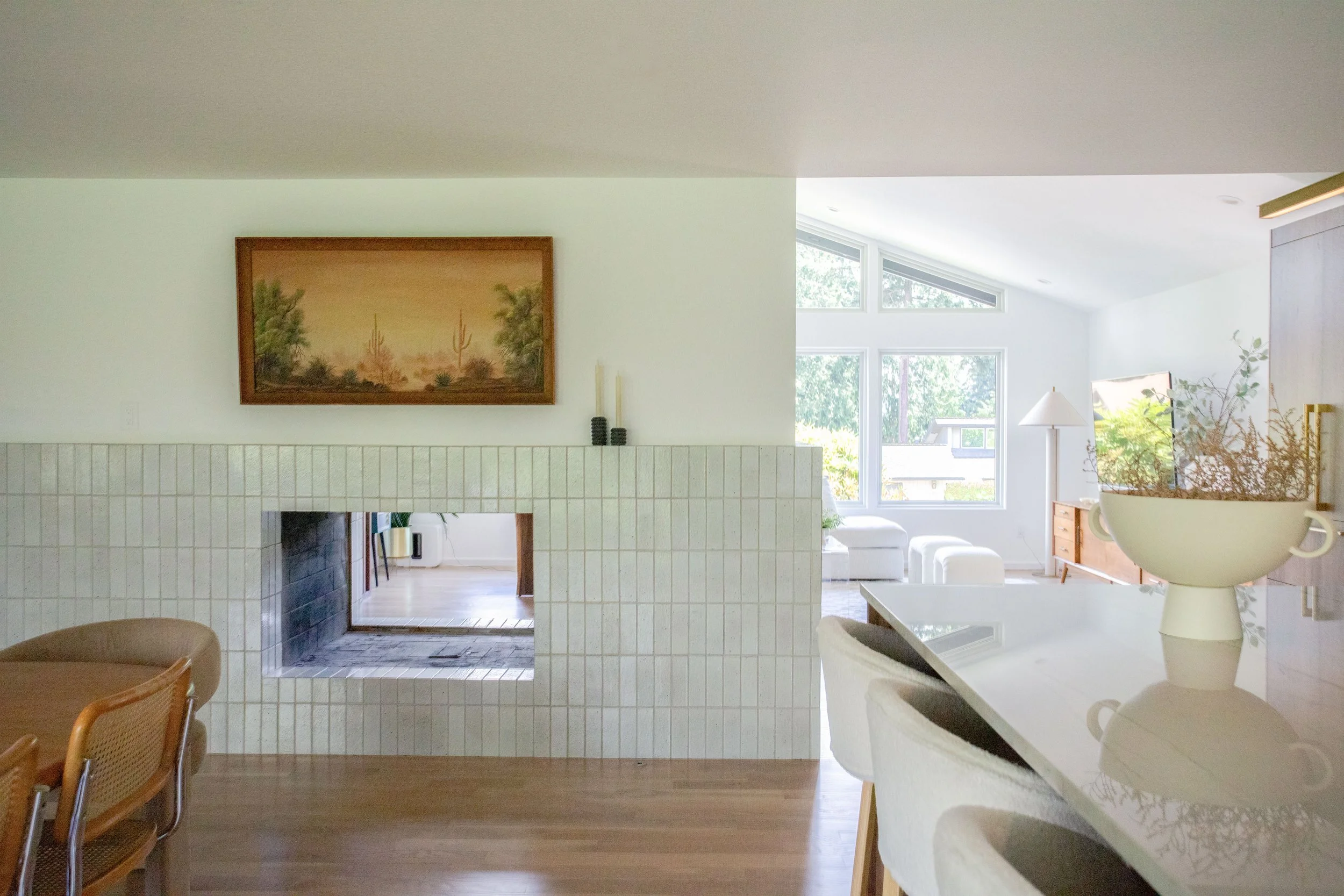 View of a bright, modern living room with large windows, white walls, a white fireplace, and a dining area with wooden chairs, decorated with plants, lamps, and wall art.