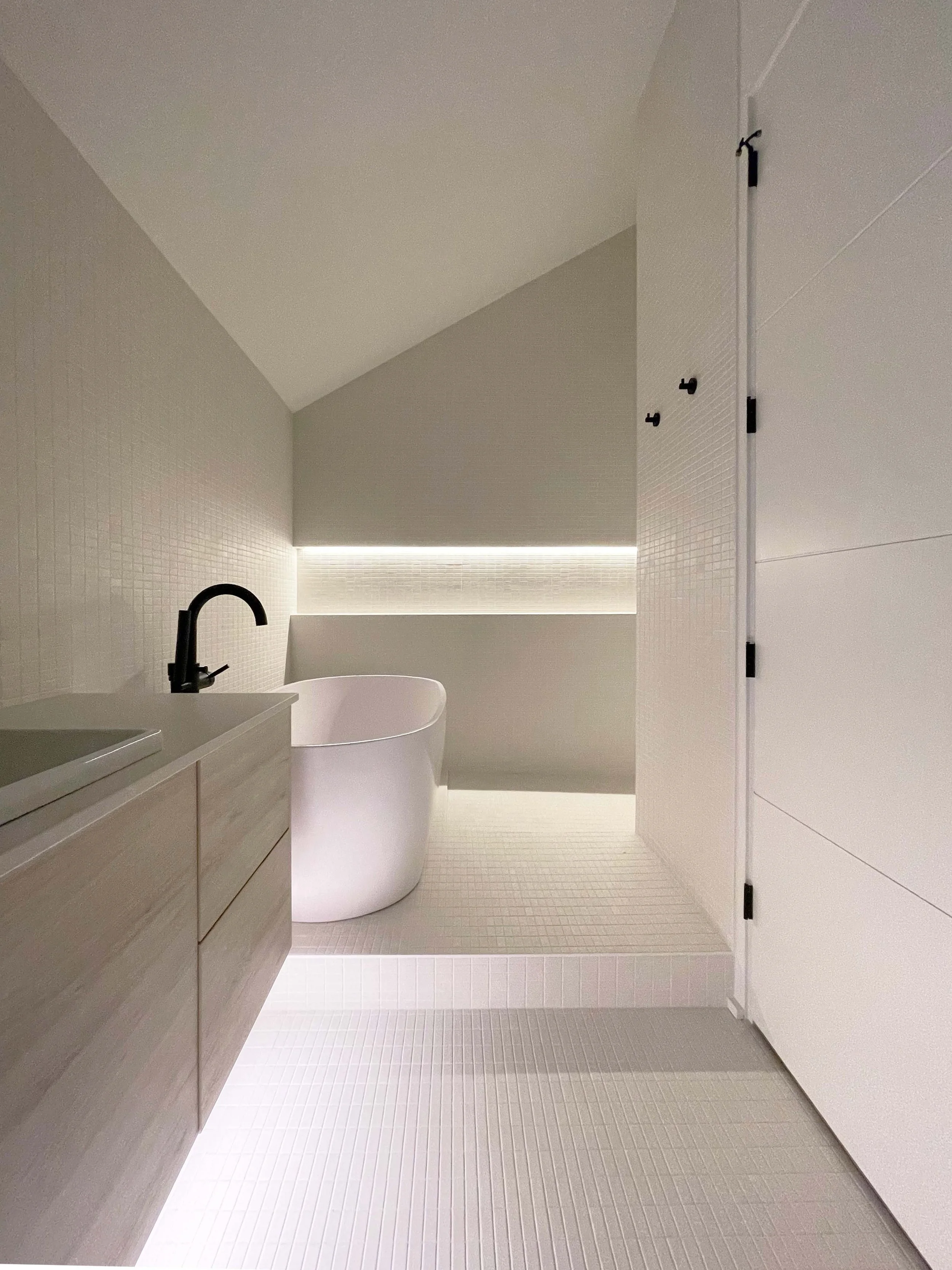 Minimalist bathroom with white walls, a white freestanding bathtub, a black faucet, a light-colored wooden vanity with drawers, and a shower area with matching white tiles and a built-in niche with lighting.