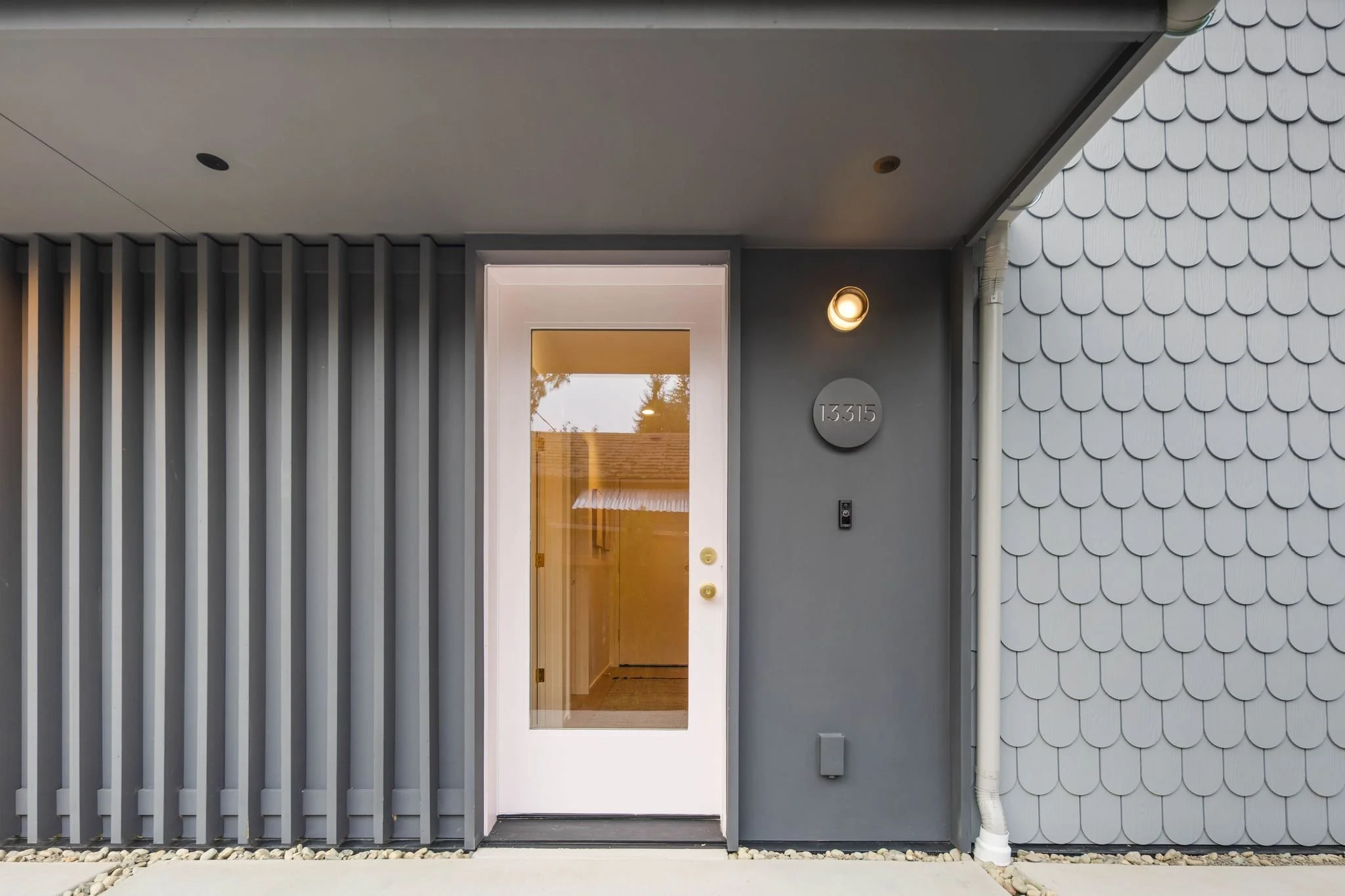 Modern house entrance with a glass door, gray siding, and decorative scalloped siding on the right, illuminated by a wall-mounted light fixture.