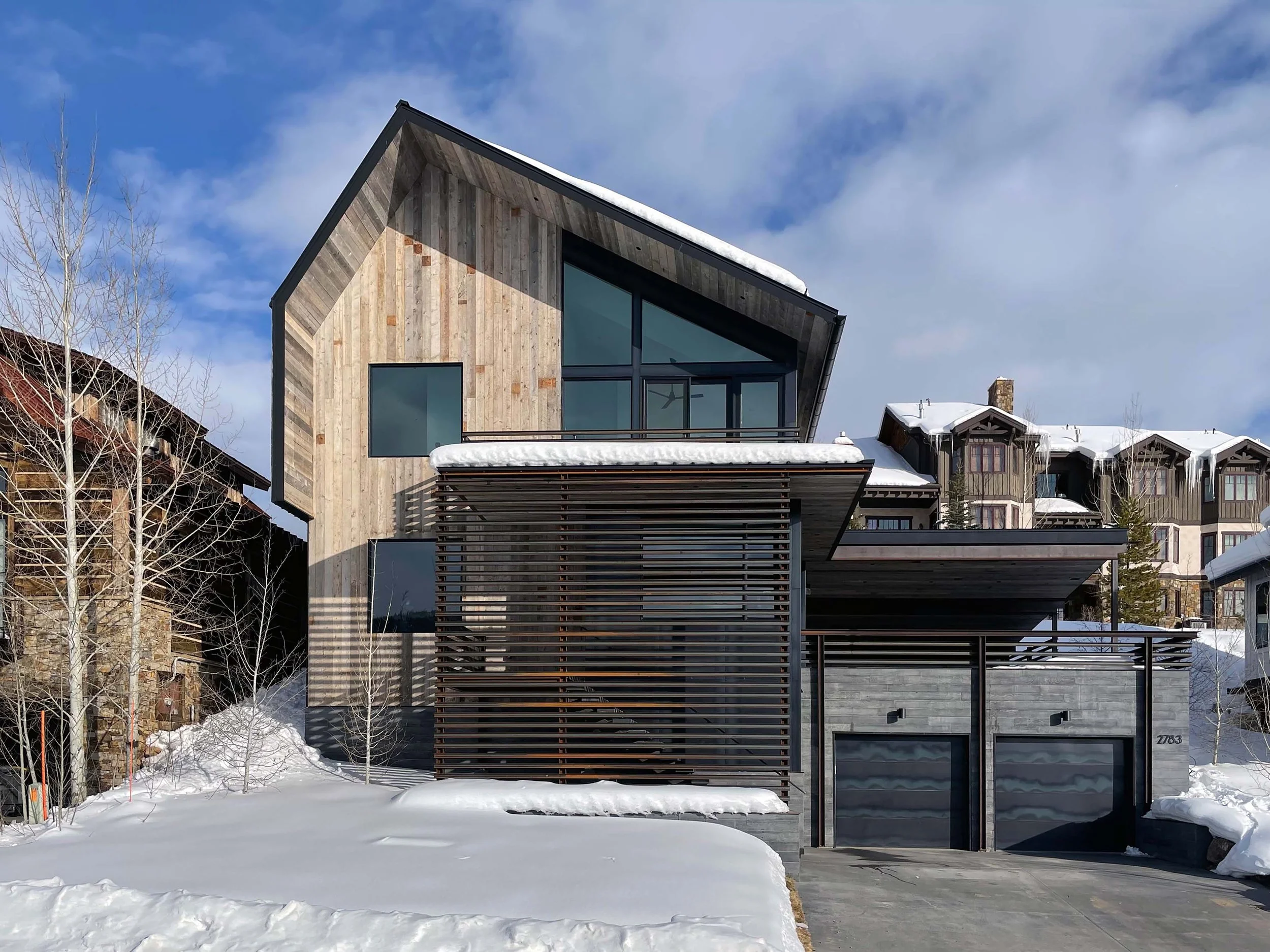 Modern house with sloped roof covered in snow, large glass windows, and wood siding in a snowy landscape with leafless trees and neighboring houses in the background.