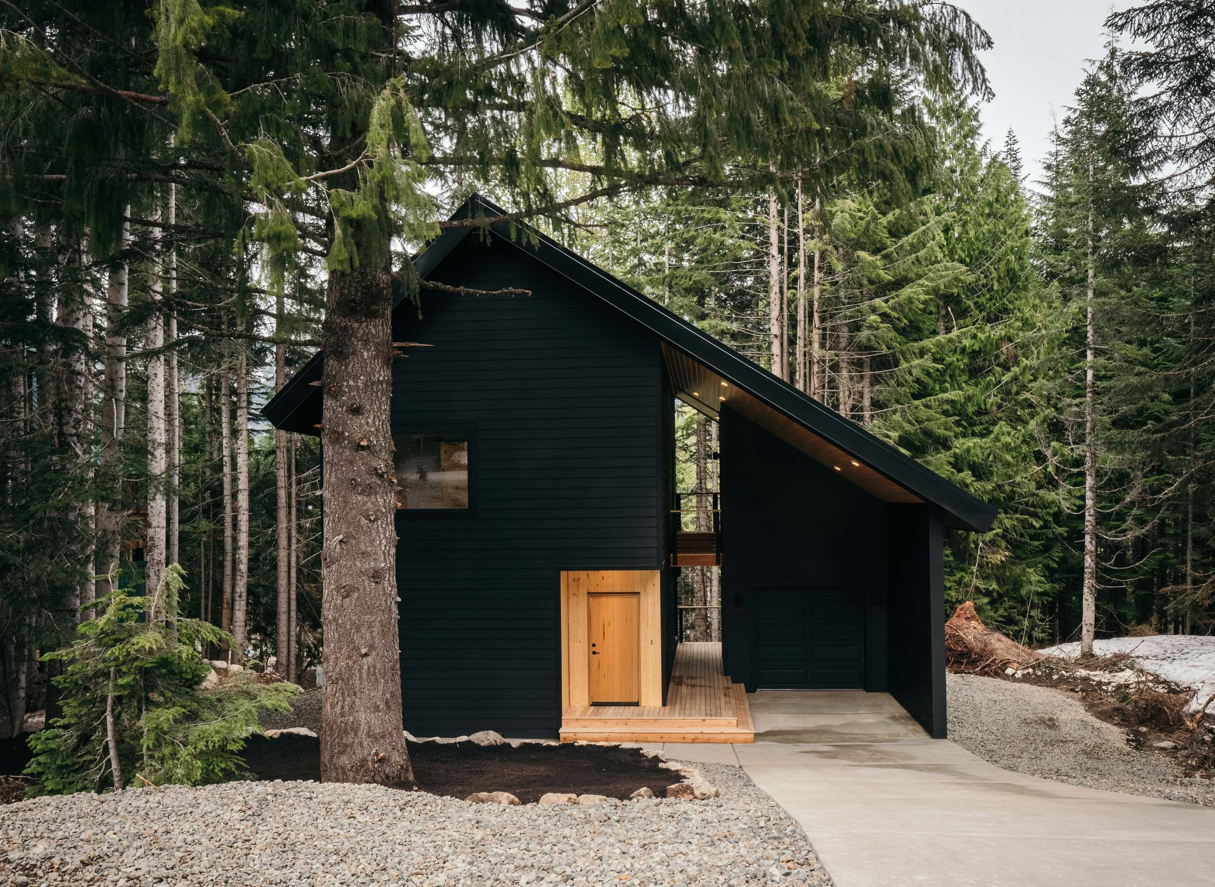 Black modern house with wooden accents, surrounded by tall green trees, in a forested area with gravel and a concrete pathway.