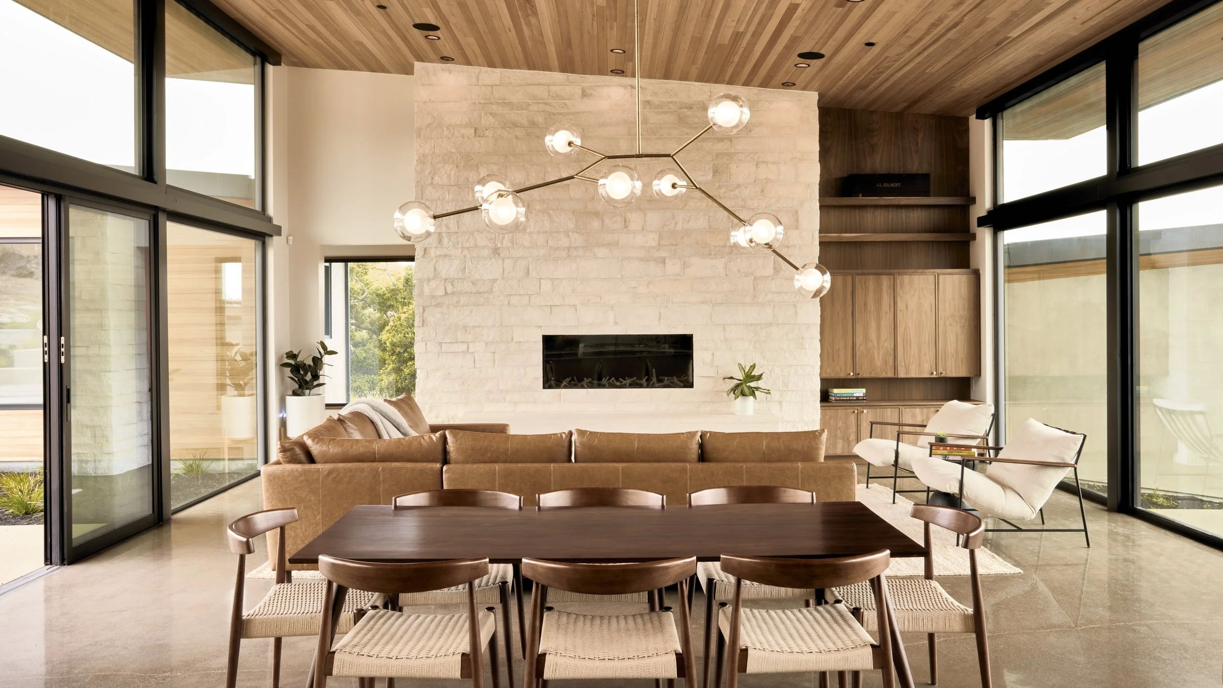 Modern living room with large windows, a white brick fireplace, a brown leather sofa, white armchairs, a wooden dining table with chairs, and a contemporary chandelier.