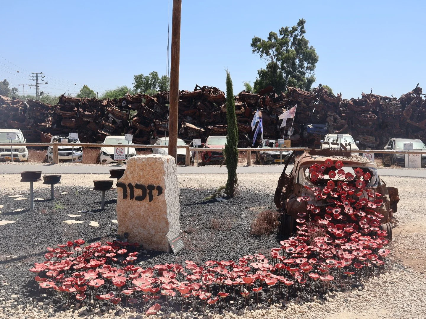A memorial site with a stone monument with Hebrew writing, surrounded by flower decorations made of red and pink poppies, with a rusty car wreckage painted with similar flowers. In the background, a fence displaying several metal plates and a large pile of rusted, crushed cars.