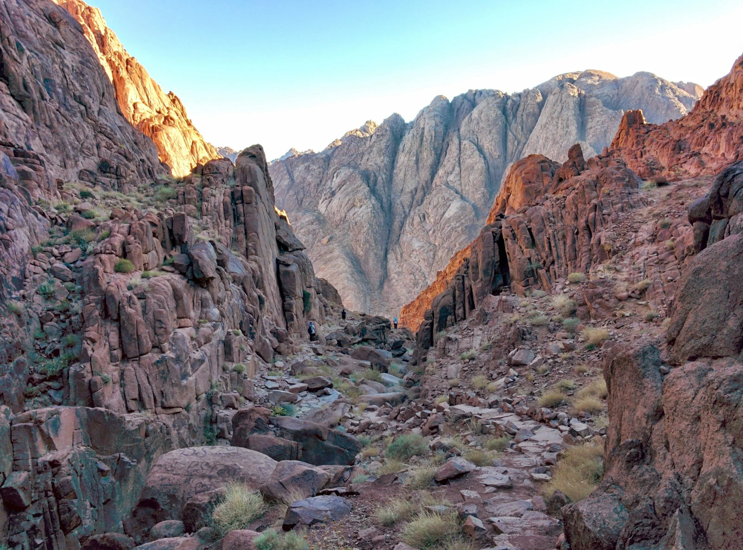 A rugged canyon with steep, rocky walls illuminated by sunlight, with hikers walking along a dirt trail at the bottom surrounded by sparse desert vegetation.